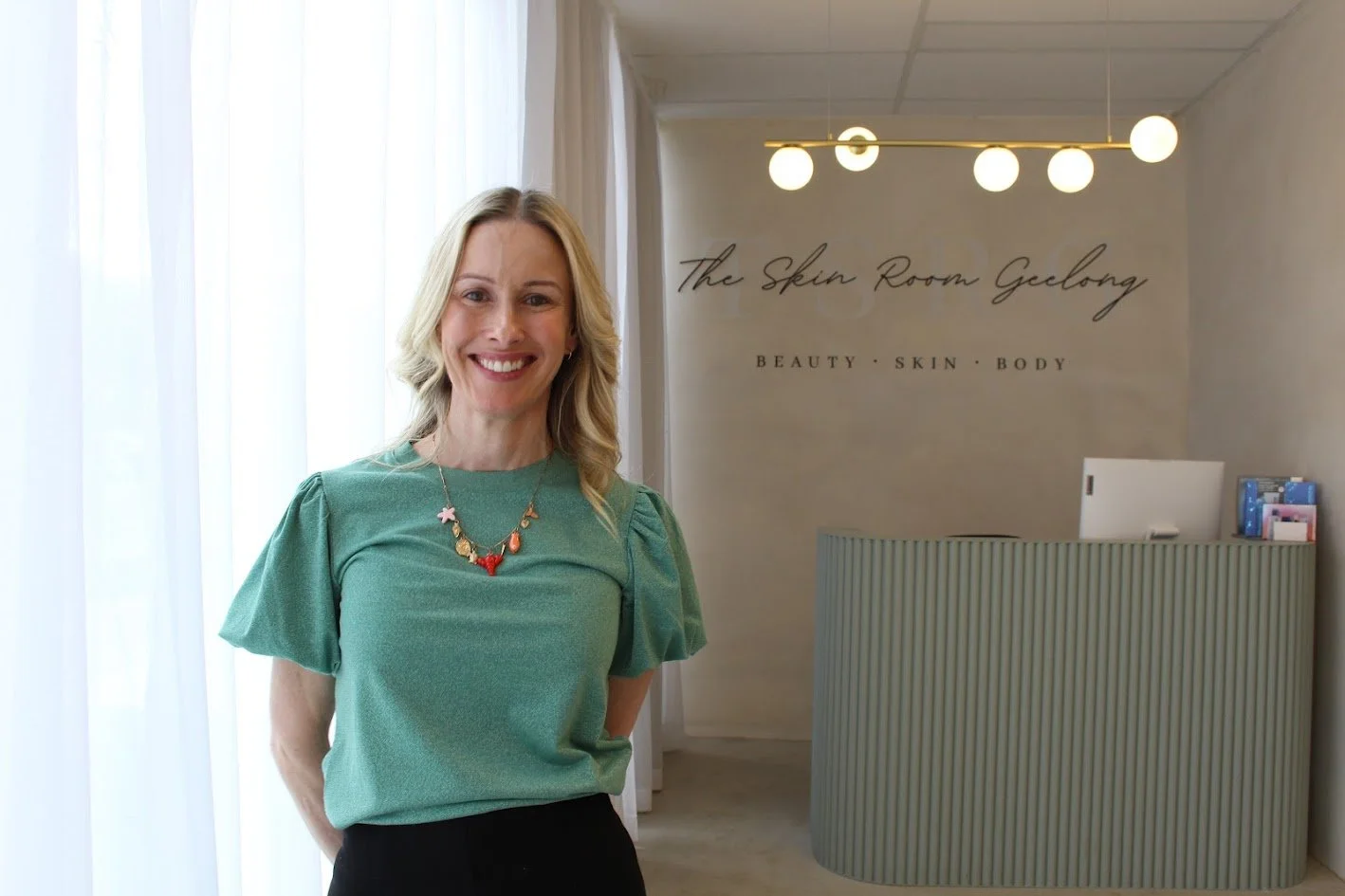 A woman with blonde hair smiling, standing in the lobby of a skincare clinic called the Skin Room Geelong, with a reception desk in the background.