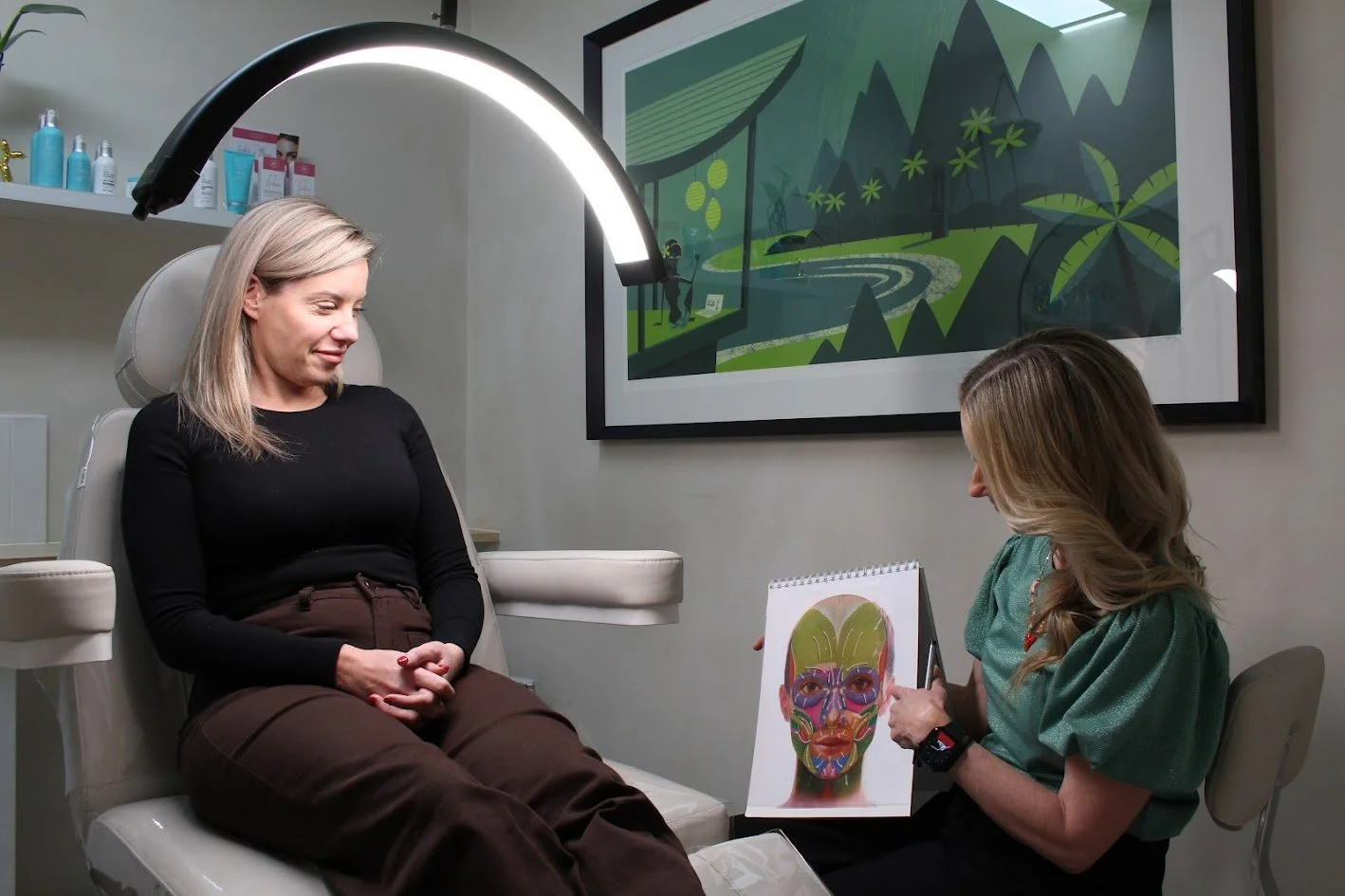 A woman sits in a medical chair with her eyes closed, during a facial consultation with a dermatologist or skincare specialist taking notes on a sketch of a face with colorful facial muscles and acupuncture points.
