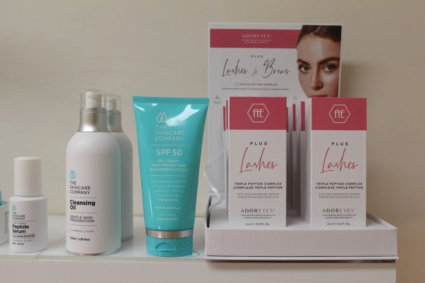 Skincare products on display including peptide serum, cleansing oil, sunscreen, and eyelash serum from Adoreyes, with promotional material in the background featuring a woman.