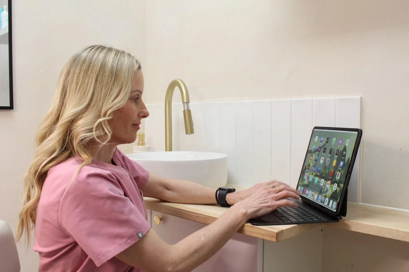 A woman with blonde hair and a pink shirt sitting at a kitchen counter using an iPad with a keyboard.