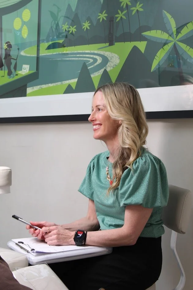 A smiling woman sitting at a table, holding a smartphone and a clipboard, with a colorful nature-themed painting on the wall behind her.