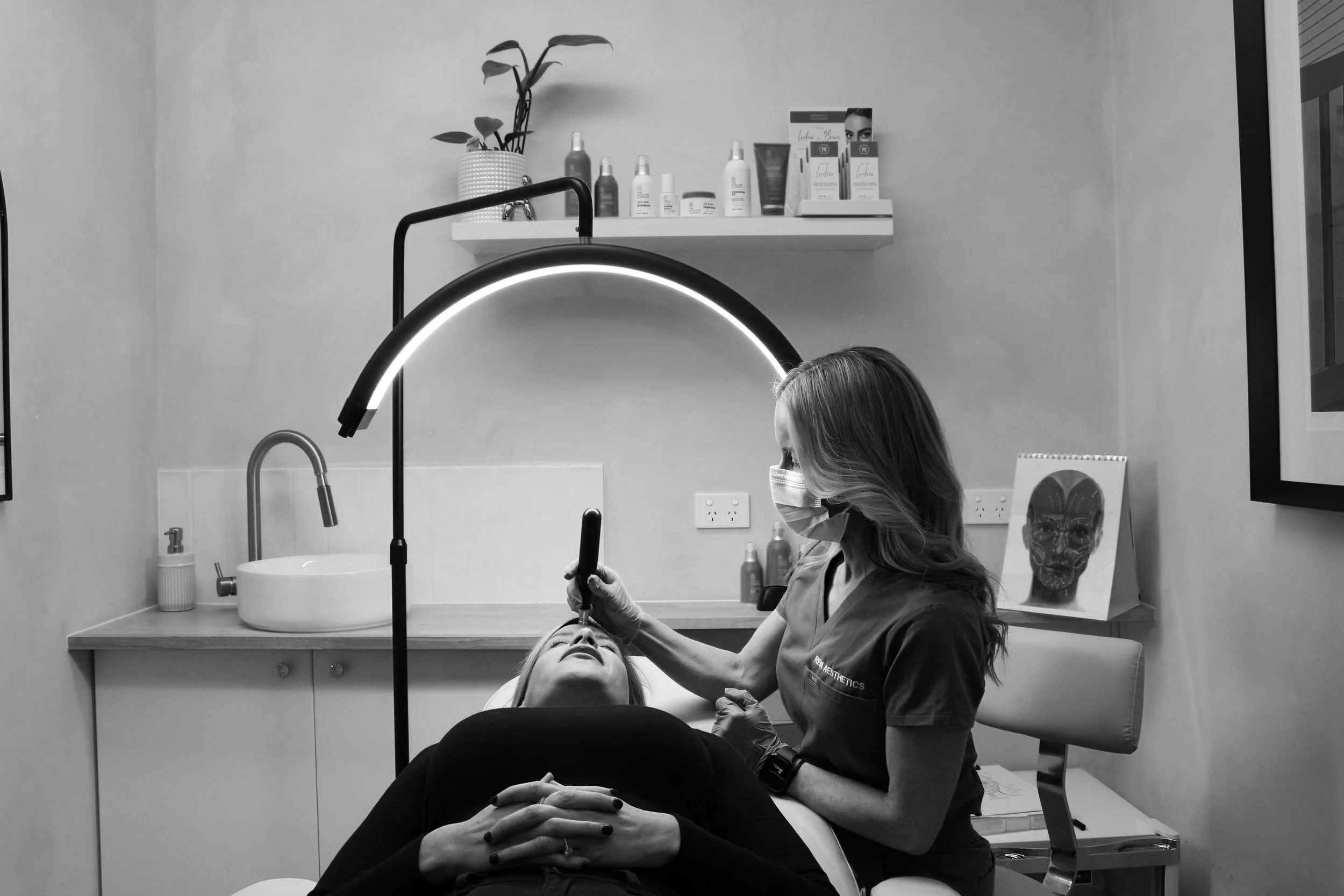 A woman receiving a facial treatment at a skincare clinic. She is lying back with eyes closed, hands folded over her stomach. A skincare professional, wearing gloves and a mask, is performing the treatment while looking at her face. The background shows shelves with skincare products, a plant, and a desk with a framed picture of a face.