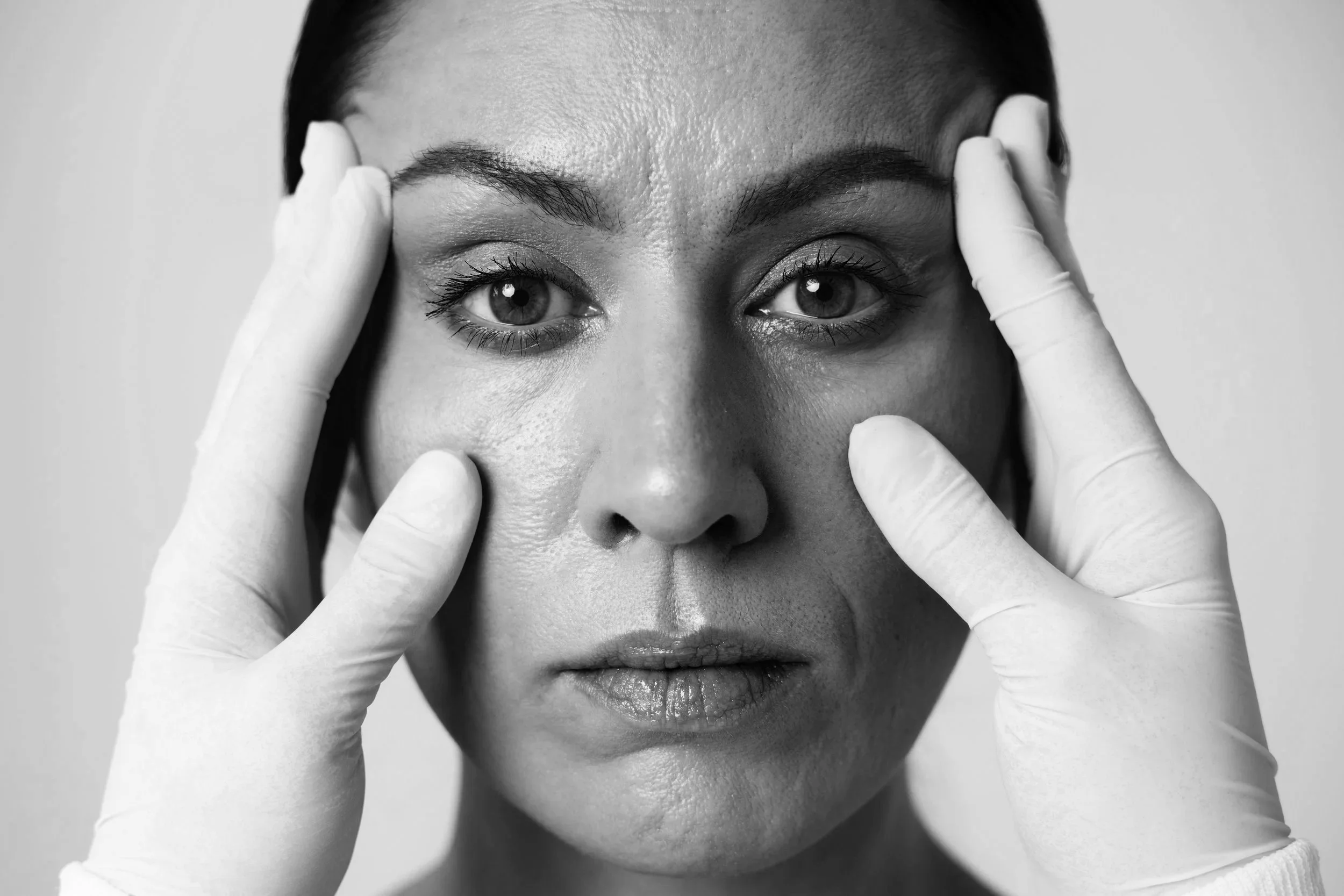 A woman with dark hair and large eyes touches her face with gloved hands, in a close-up black and white portrait.