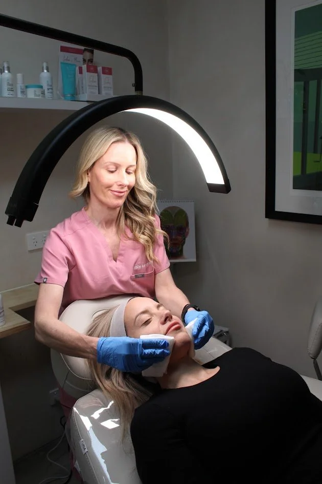 A woman lying down on a treatment chair with her eyes closed, receiving a skincare facial from a female skincare specialist wearing pink scrubs and blue gloves. The specialist is applying a treatment under a large, illuminated lamp.