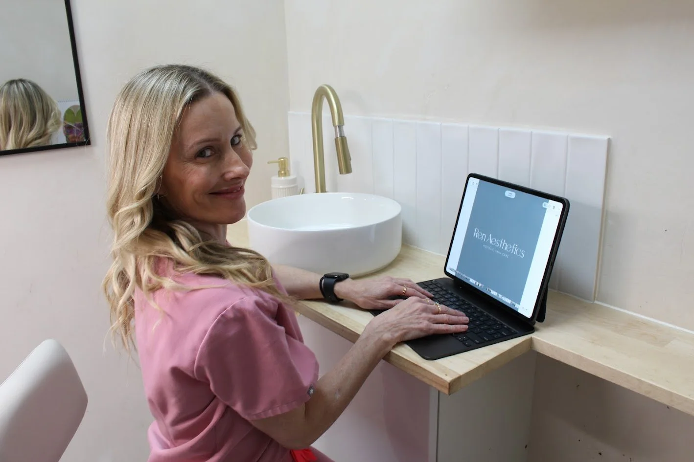 Woman with blonde hair in pink shirt sitting at a wooden counter with a sink, using a tablet with a keyboard in a modern setting.