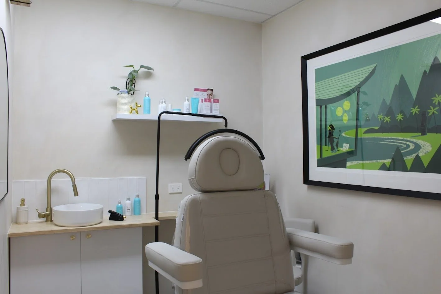A medical examination room with a beige chair in front of a large framed landscape artwork, a white sink with a gold faucet, and shelves holding skincare products, a potted plant, and a small mirror.