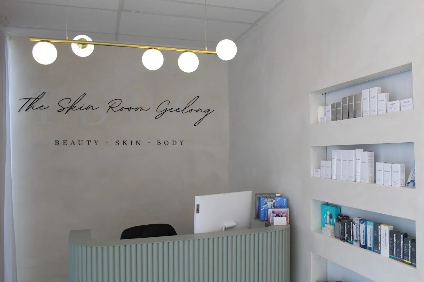 Reception area with wall sign reading 'The Skin Room Geelong' and tagline 'Beauty, Skin, Body'. White curved reception desk, black chair, two recessed shelves with skincare products, modern round ceiling light fixture with five globe bulbs.