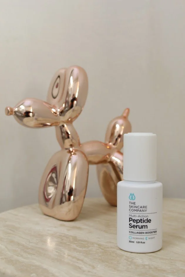 A white bottle of The Skincare Company Peptide Serum with a small blue water droplet logo on the label, placed on a wooden surface, with a metallic balloon sculpture of a dog in the background.