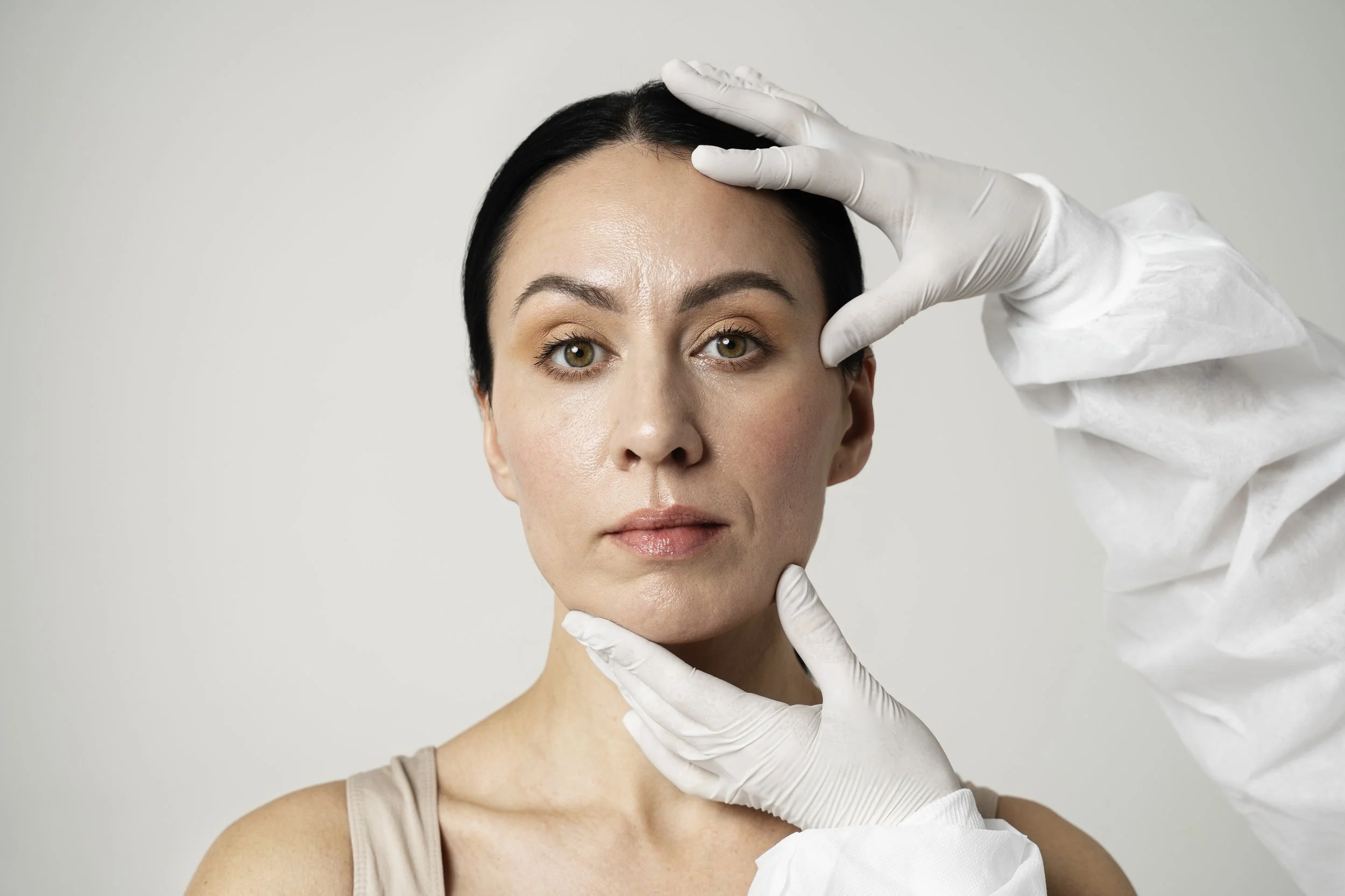A woman with short dark hair and light brown eyes has her face examined by a person wearing white gloves and a white coat, on a neutral grey background.