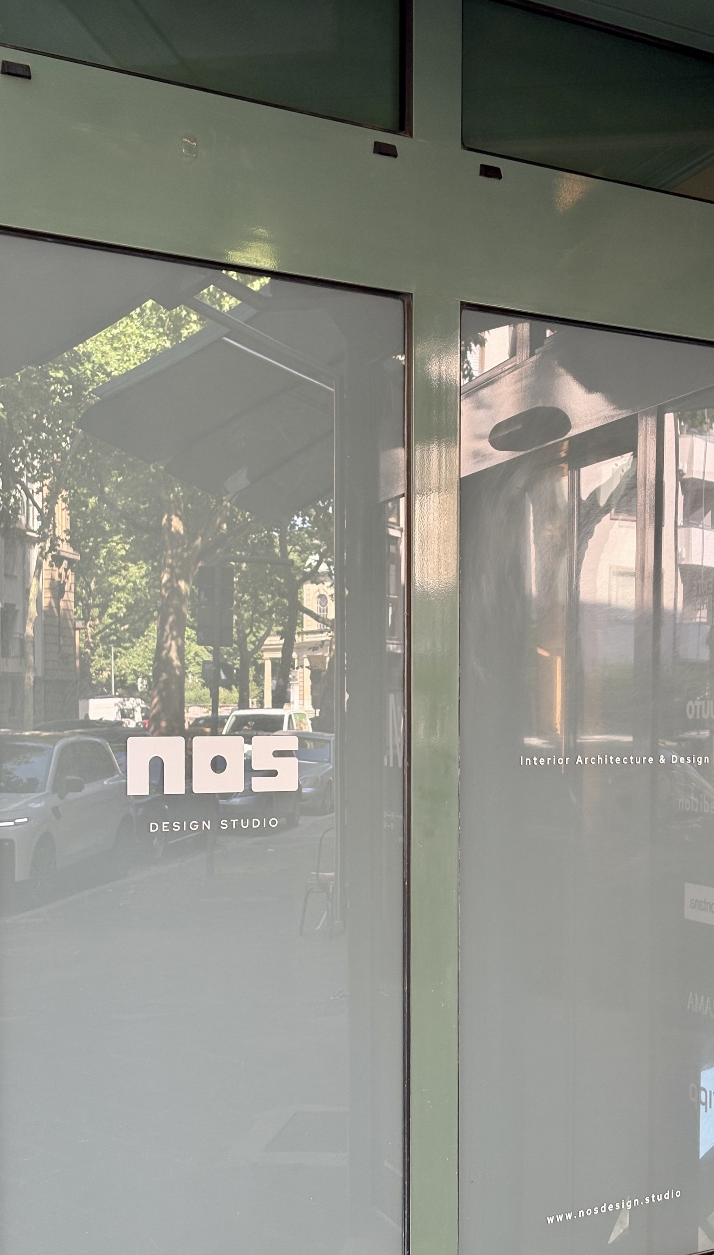 Fensterscheibe mit einem Logo und Text, das auf ein Designstudio namens Nos Design Studio hinweist. Es ist die Elisabethstrasse in Mannheim mit Autos und Bäumen sichtbar.