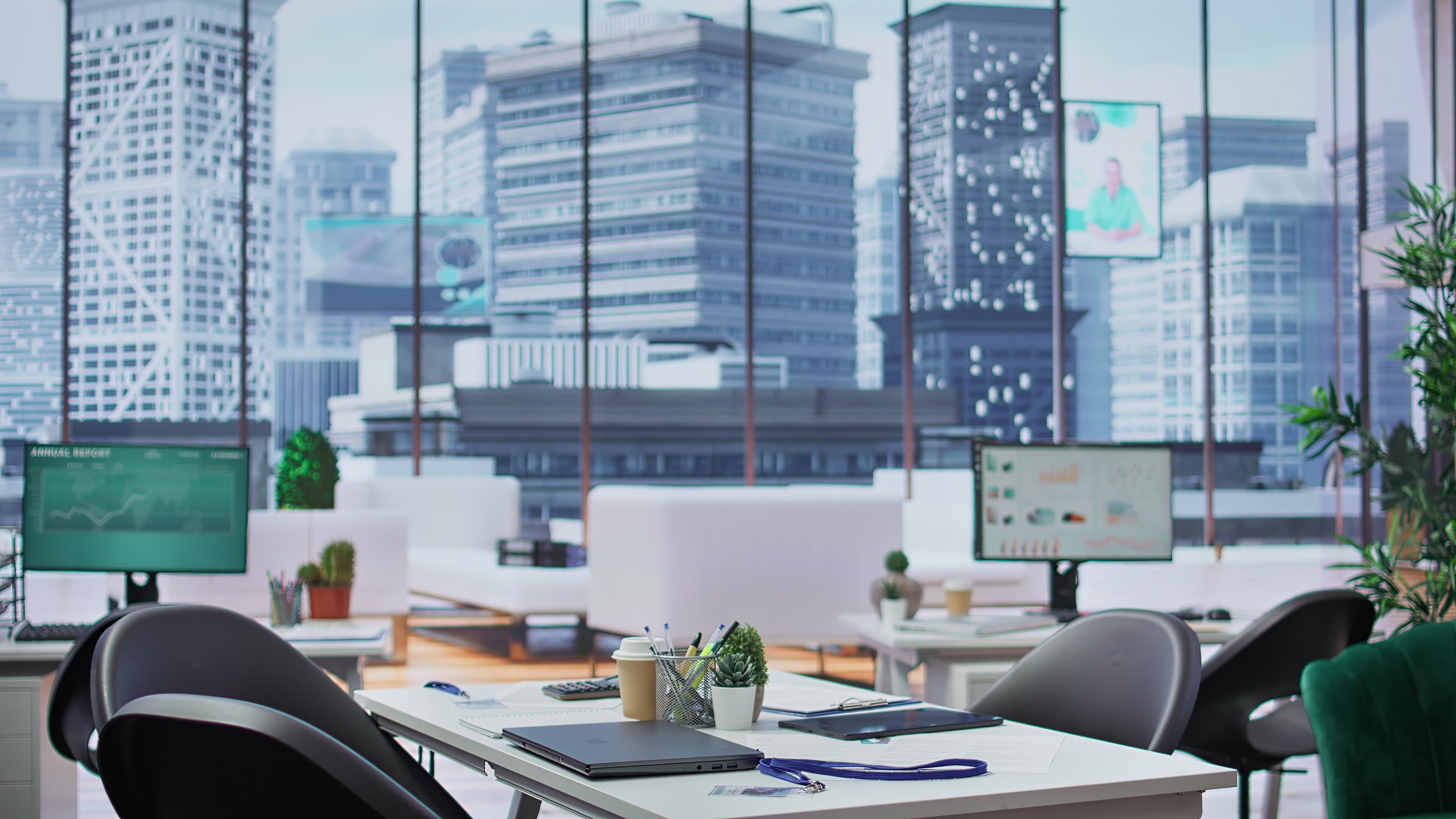 Modern office with large windows showing a cityscape of tall buildings. Inside, there are white desks with laptops, office supplies, plants, and chairs.