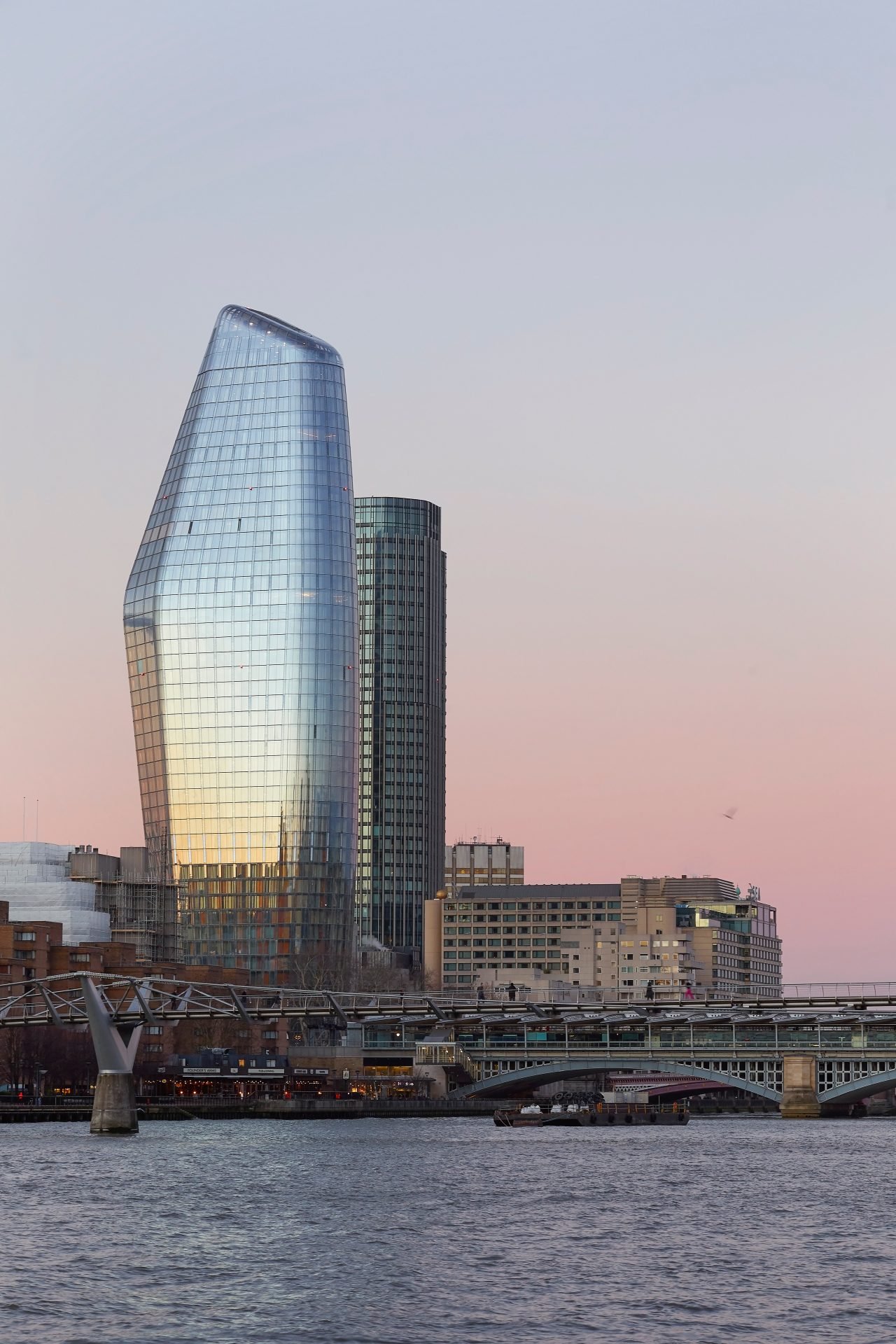 Photograph of a modern skyscraper with a curved, reflective glass facade, set against a pastel-colored sky during dusk, with surrounding buildings and a river in the foreground.