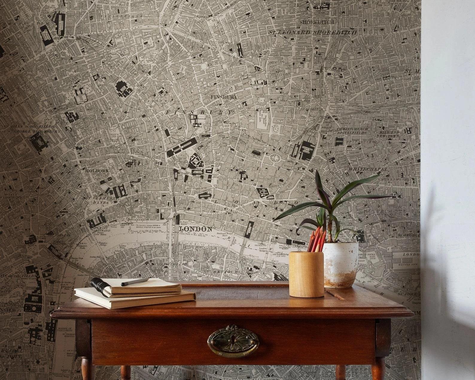 A wooden desk with a stack of notebooks and a pen, a potted plant with long leaves in a small pot, and a large wall map of London, England in the background.