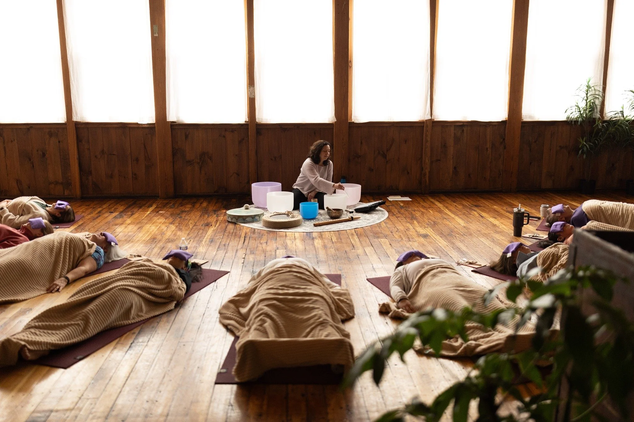 A woman leading a group meditation session indoors with wooden walls, large curtains, and a potted plant, all seated on the floor with their eyes closed and wearing beige blankets.
