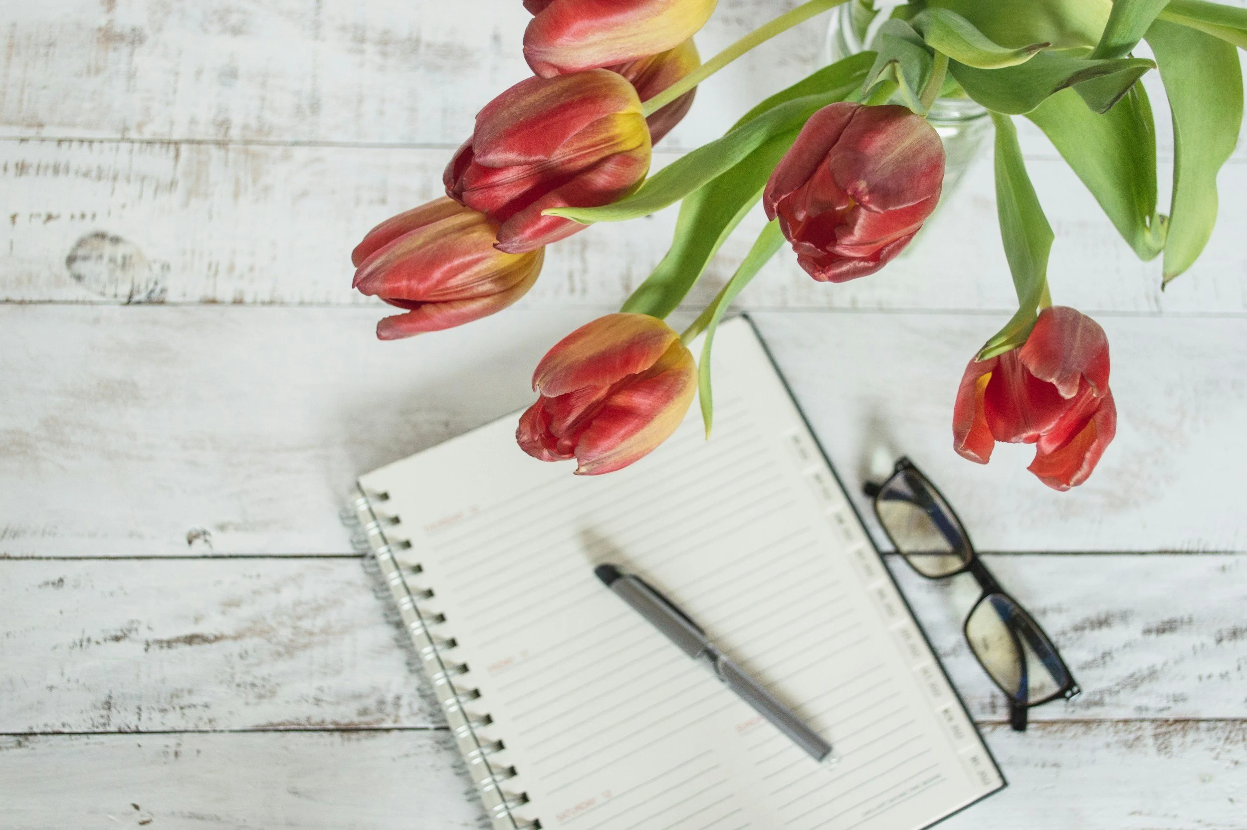 A bouquet of red and yellow tulips in a glass vase on a white wooden surface with an open planner, a pen, and a pair of glasses nearby.