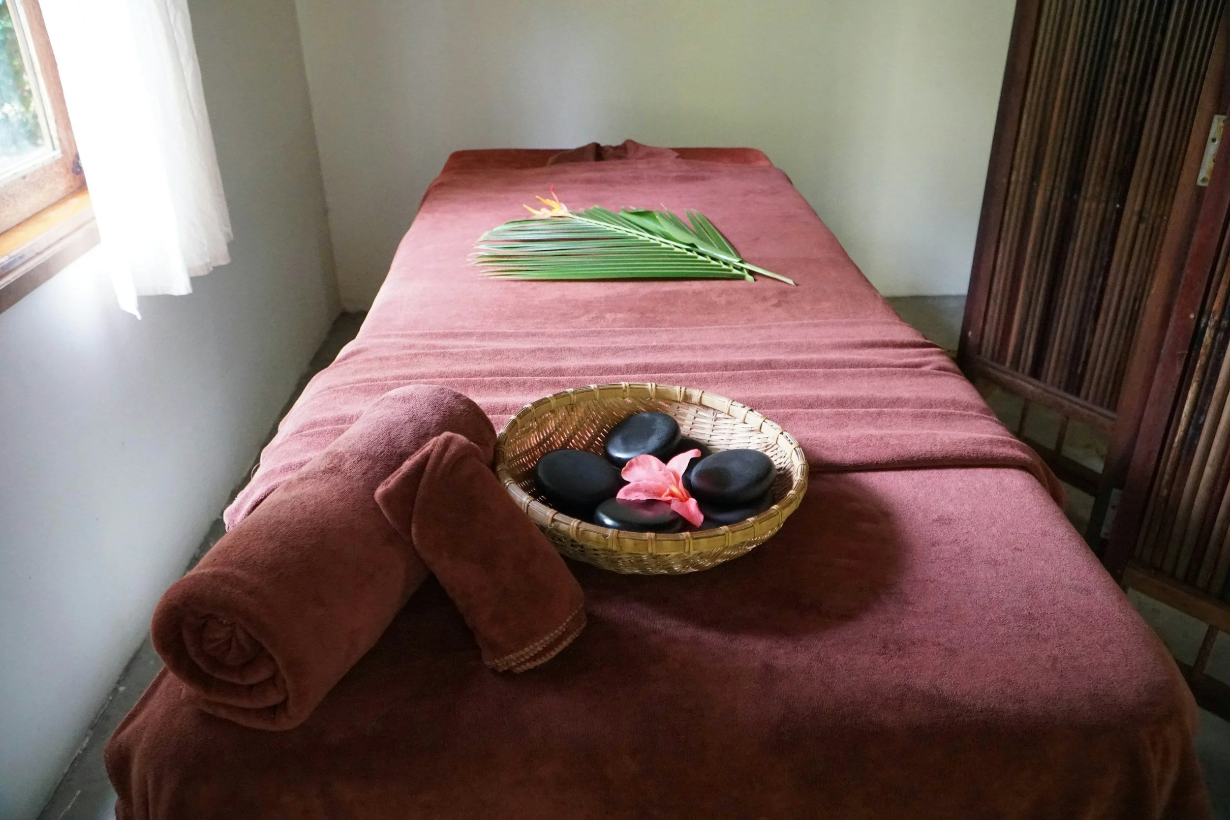 A massage table covered with a pink towel, with a green palm leaf, a basket containing black stones, and a pink flower on top of it inside a room with a window and white curtains.