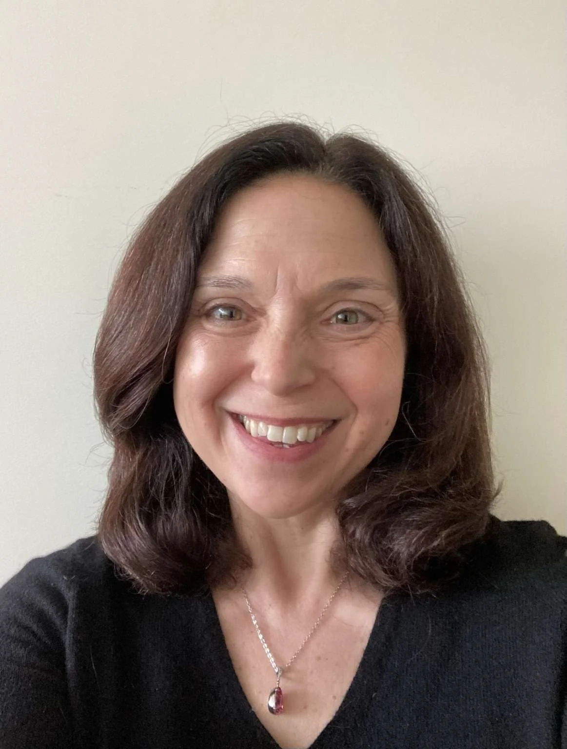 A smiling woman with shoulder-length dark brown hair, wearing a black top and a silver necklace with a red pendant, standing against a plain light-colored wall.