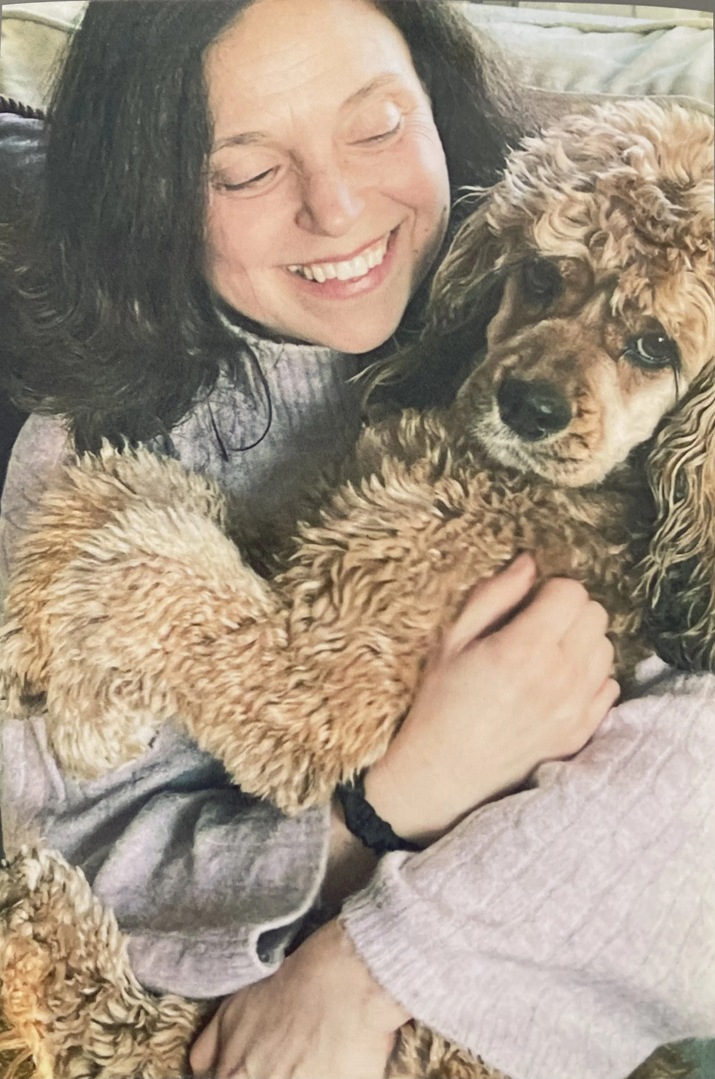 A woman with curly dark hair smiling while cuddling a golden-brown poodle dog with curly fur.