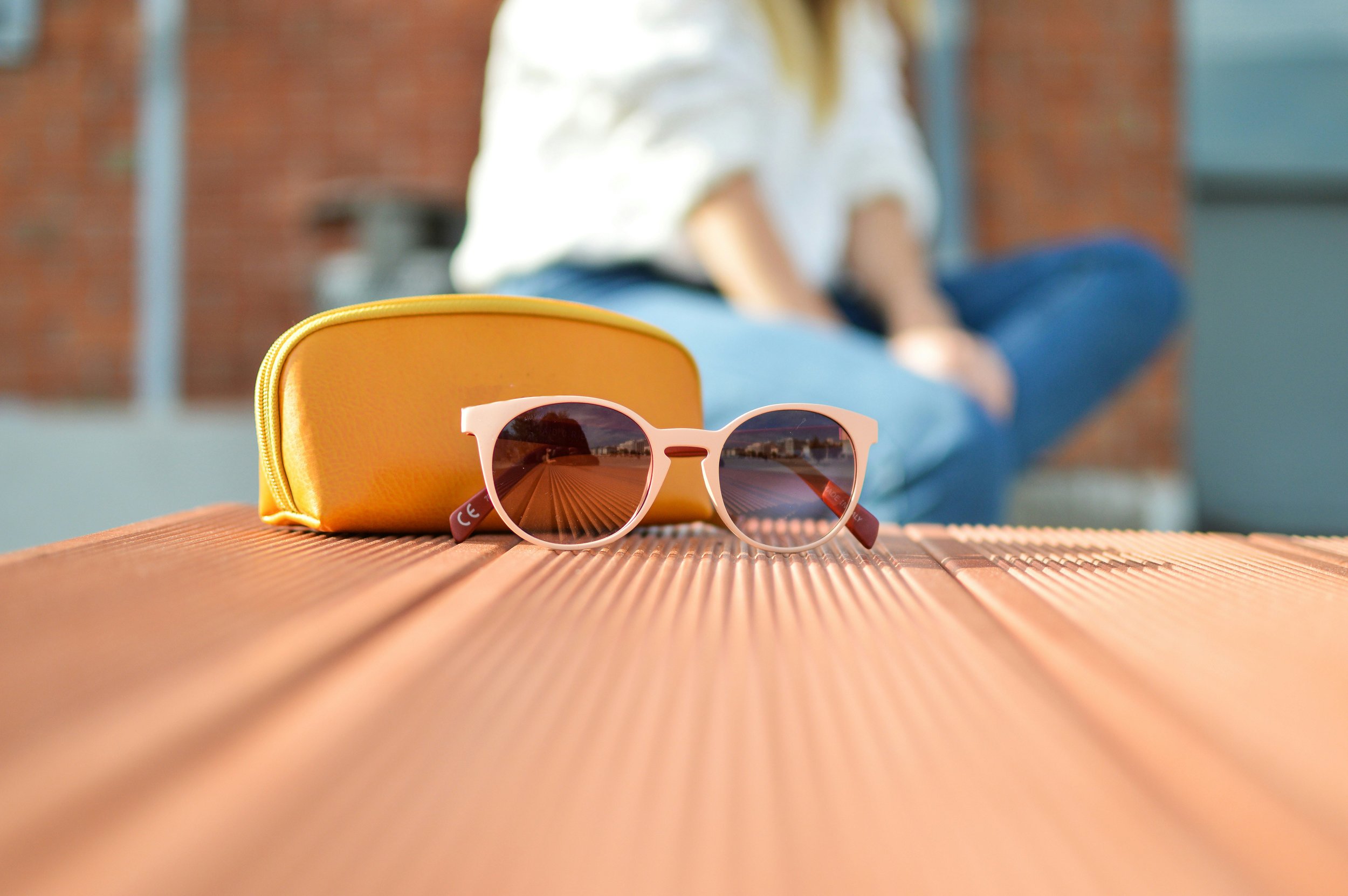 Pink sunglasses with a reflection of a wooden pier, yellow case, and a blurred person sitting in the background.