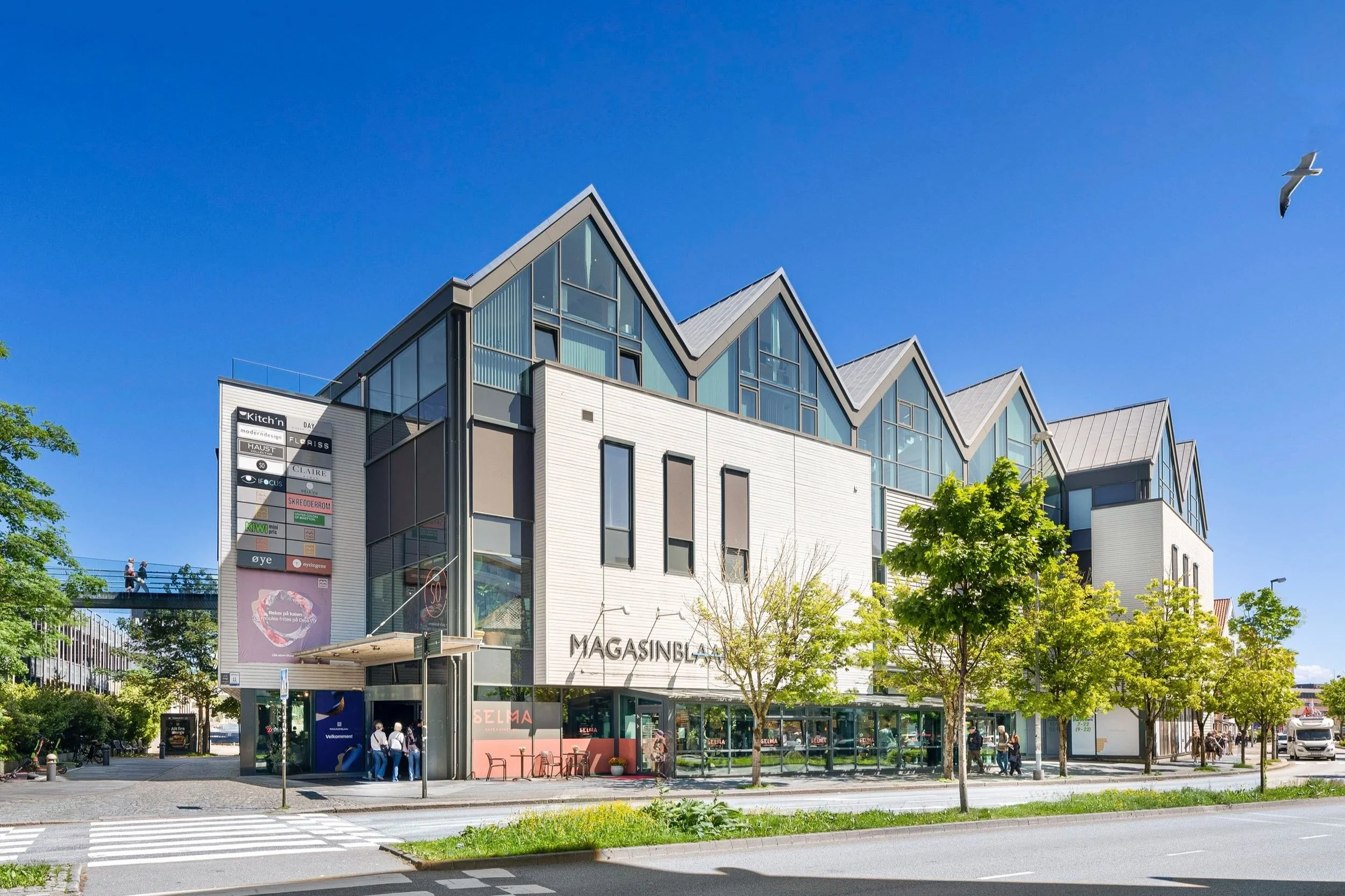 Modern multi-story shopping mall with glass facade, trees, and people outside on a sunny day.