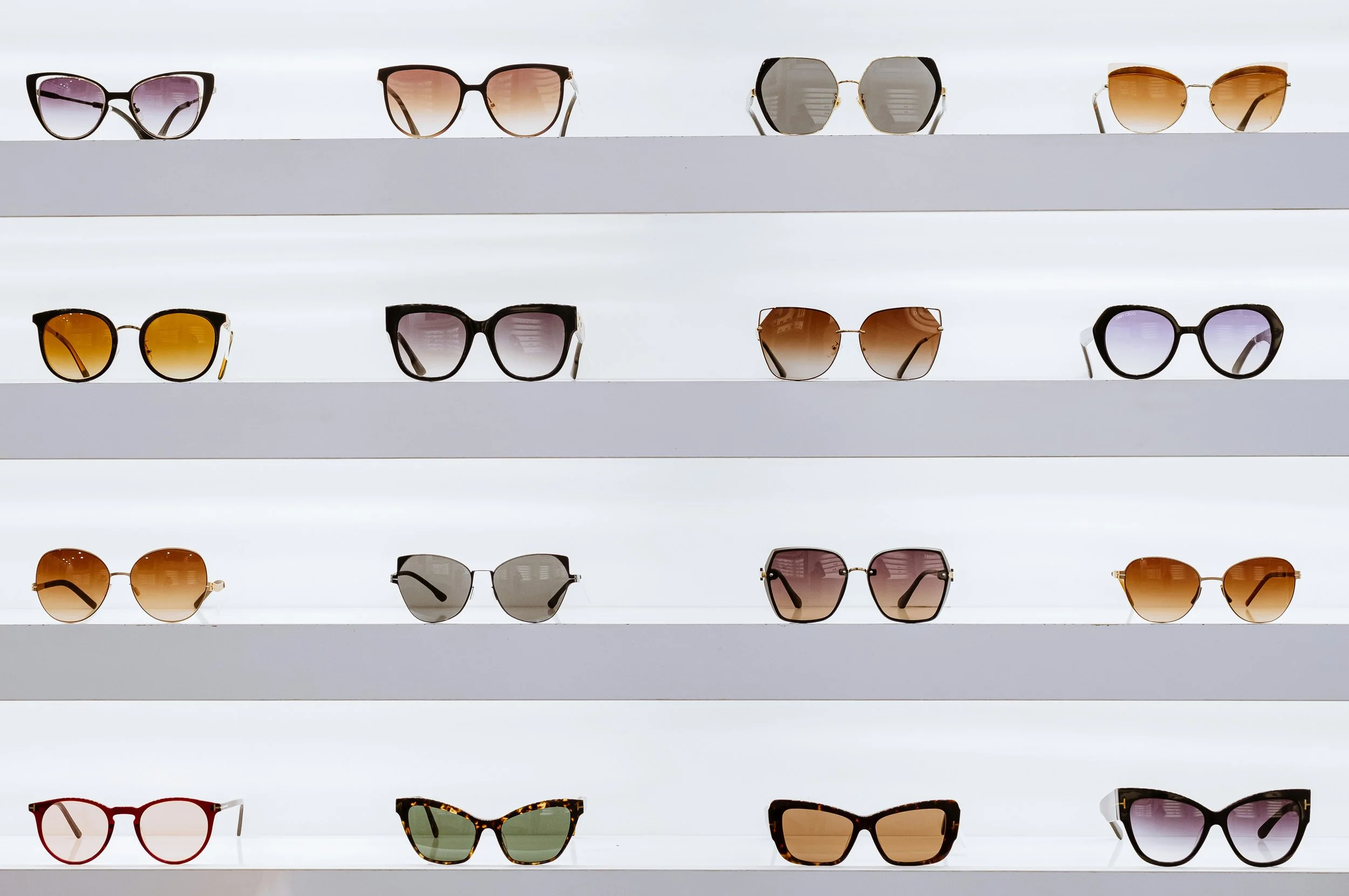 Multiple pairs of sunglasses with various styles and colors displayed on white shelves.