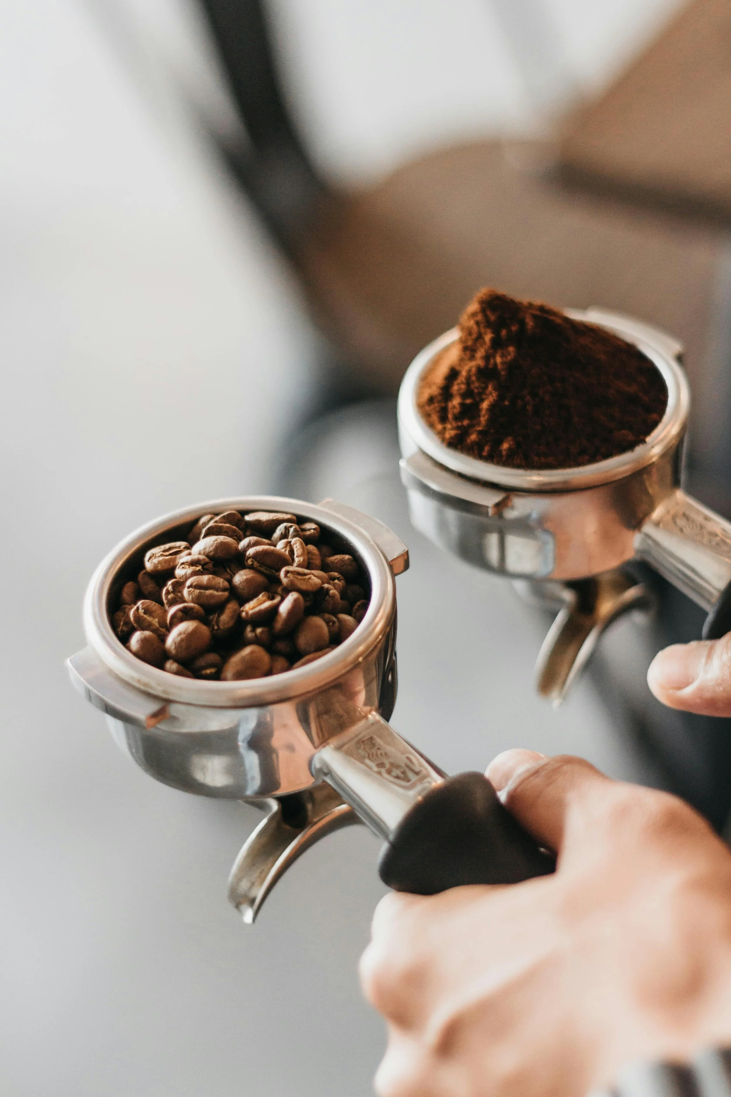 Close-up view of a portafilter filled with coffee beans and another filled with ground coffee, being held by a person.