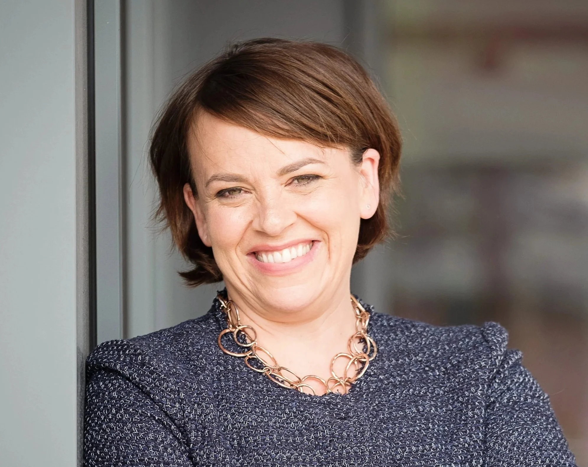 A woman with short brown hair and a big smile, wearing a dark patterned blazer and a large gold necklace, standing in front of a grey background.