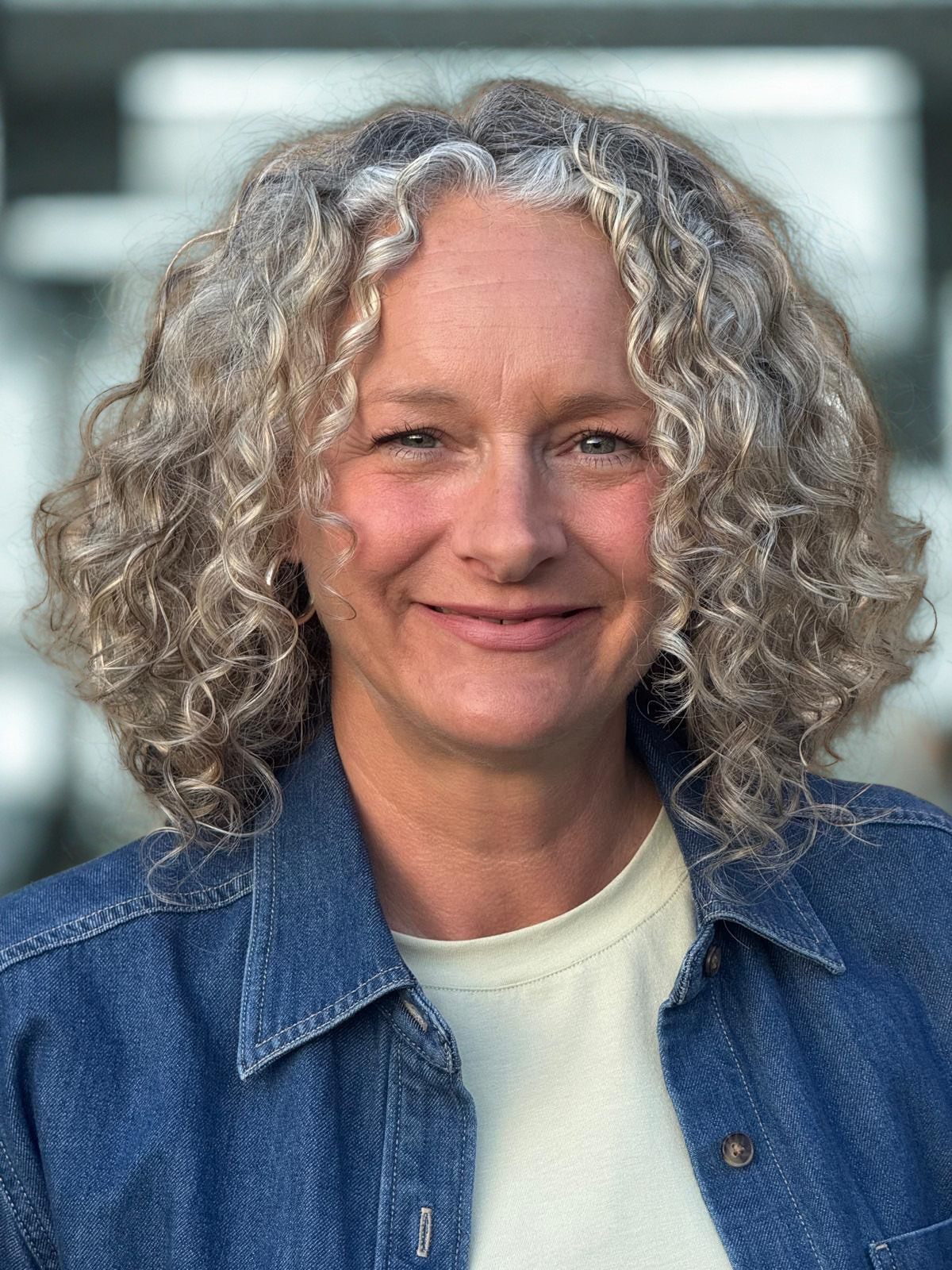 A woman with curly, gray hair smiling, wearing a denim jacket over a light-colored shirt, standing in an indoor setting with a blurred background.