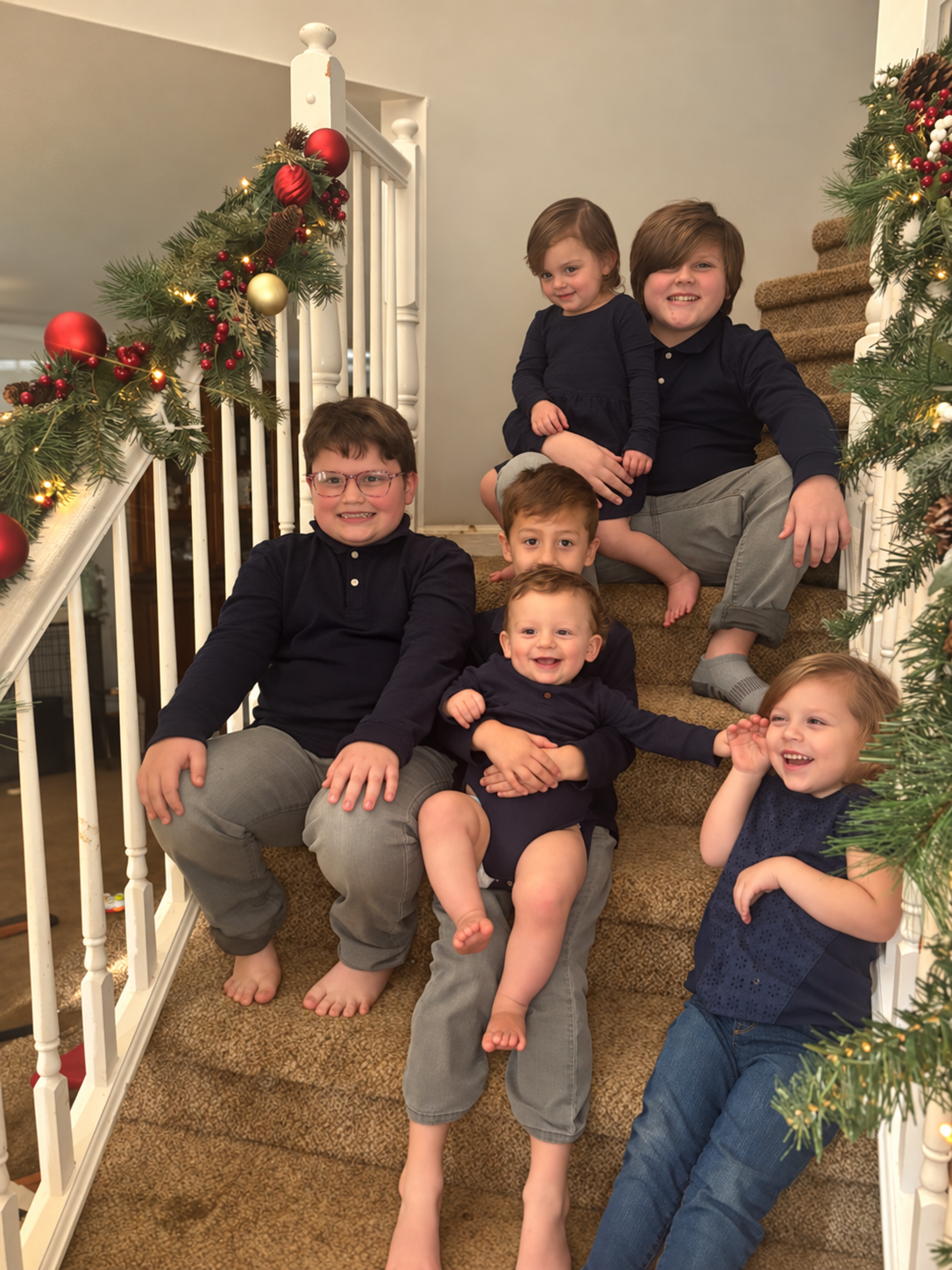 A group of children sitting on a decorated staircase with Christmas garland, smiling and posing for a photo.