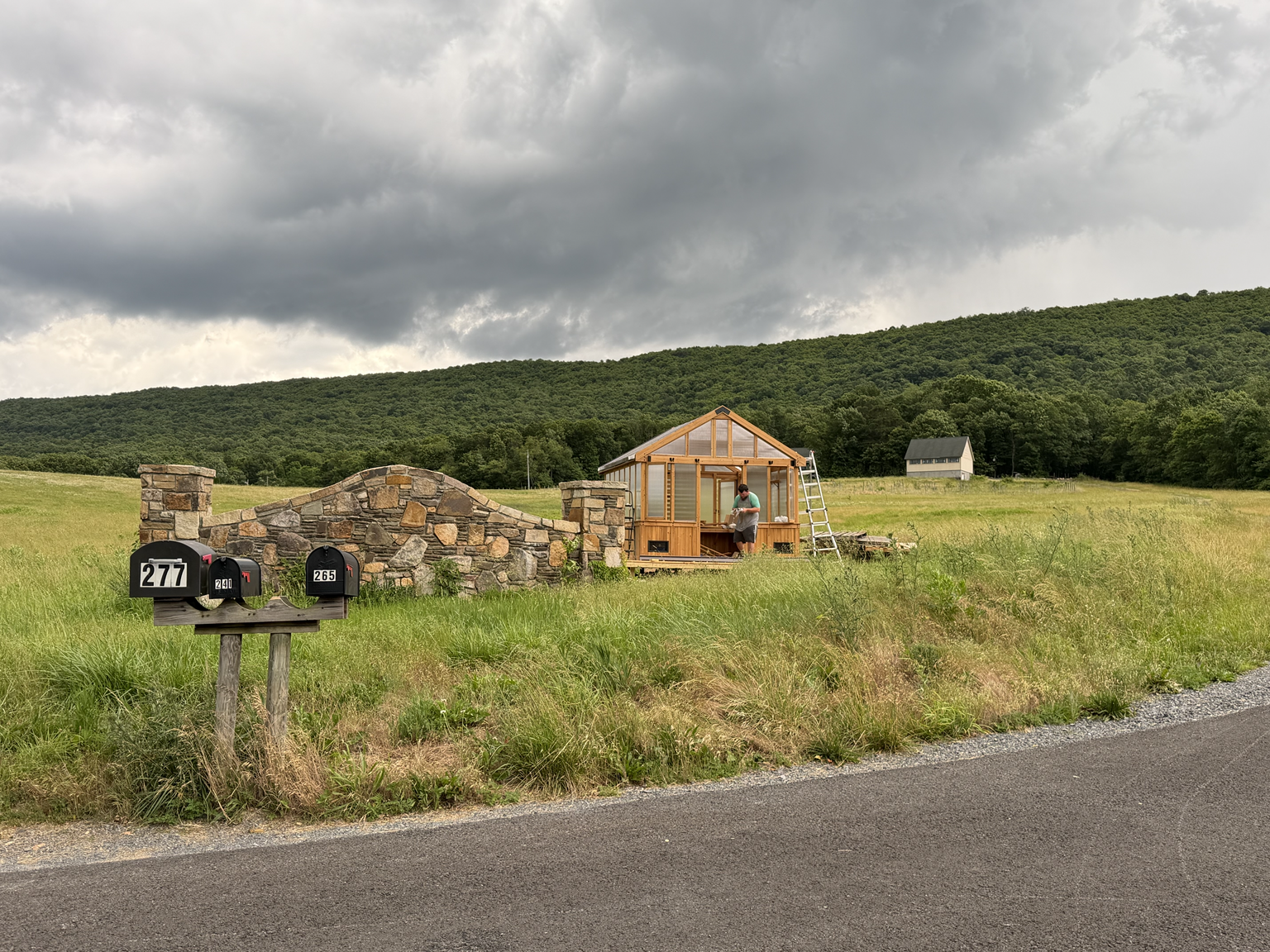 A person building a small wooden greenhouse on a grassy field with a stone wall and mailbox in the foreground, and mountains with dark clouds in the background.
