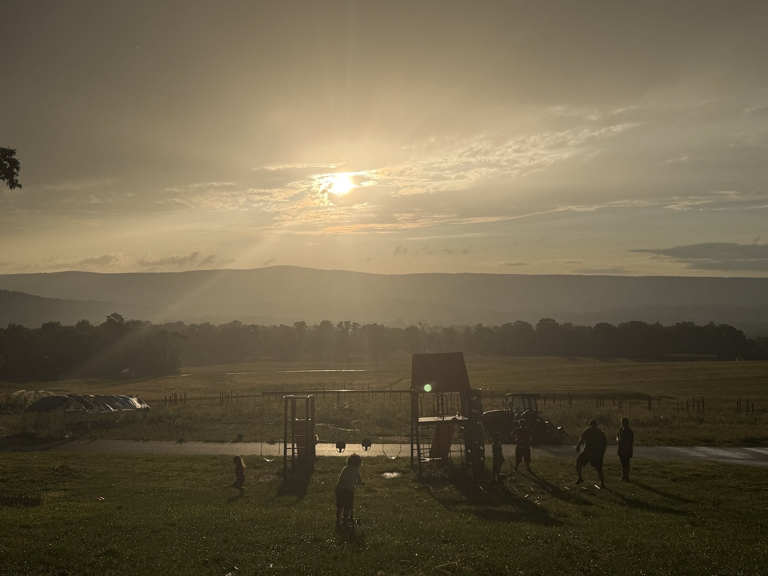 Children and adults playing and standing near playground equipment outdoors during sunset, with fields and mountains in the background.