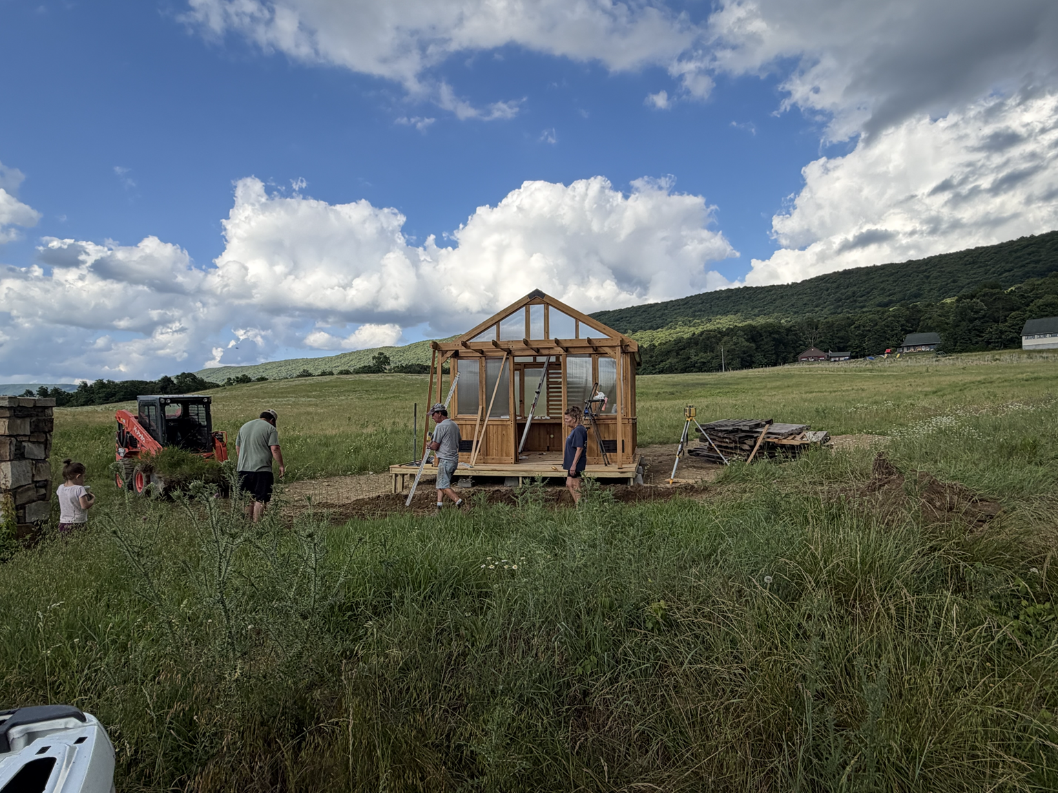 People building a wooden greenhouse in an open field on a sunny day with a partly cloudy sky.