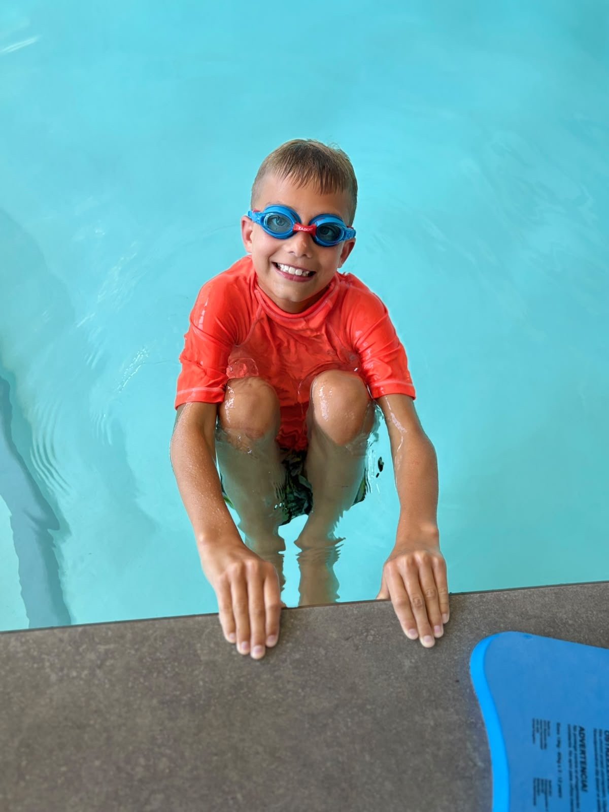 A young boy wearing swimming goggles and a red swim shirt, smiling while sitting on the edge of a swimming pool with his arms resting on the poolside.