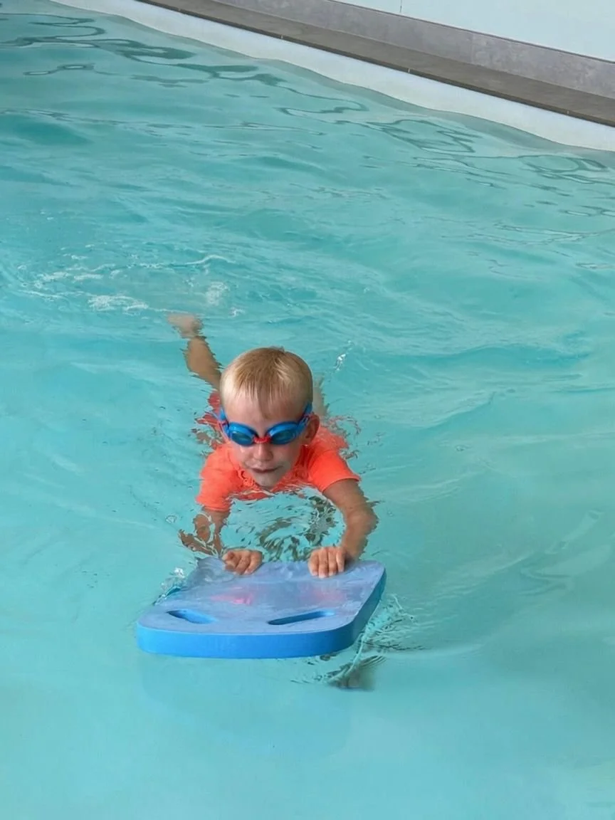A young boy swimming in a pool wearing orange swimwear and blue goggles, holding a blue kickboard.