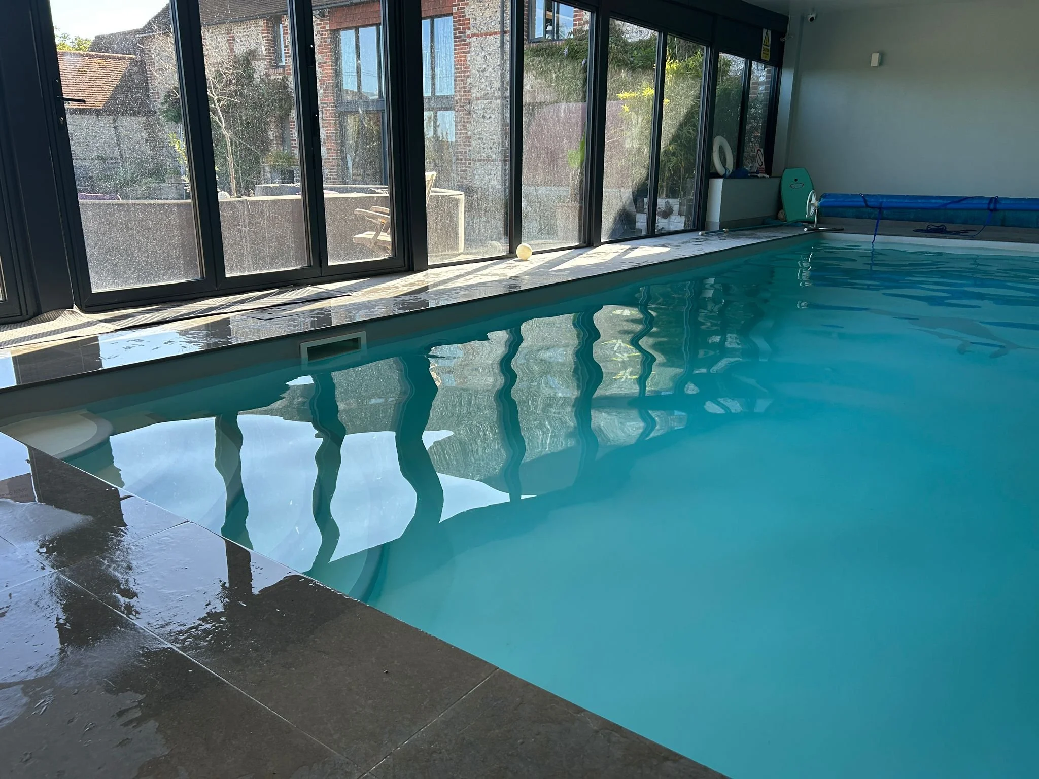 Indoor swimming pool with large glass sliding doors overlooking outdoor buildings and trees, with pool accessories on the side.