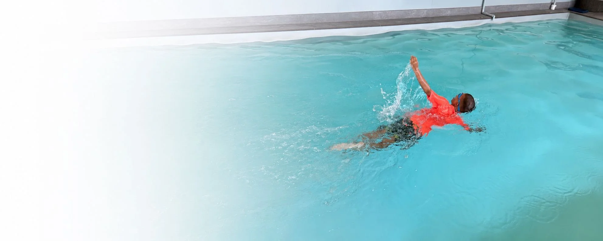 Child swimming in an indoor pool, wearing red swim shirt and blue goggles