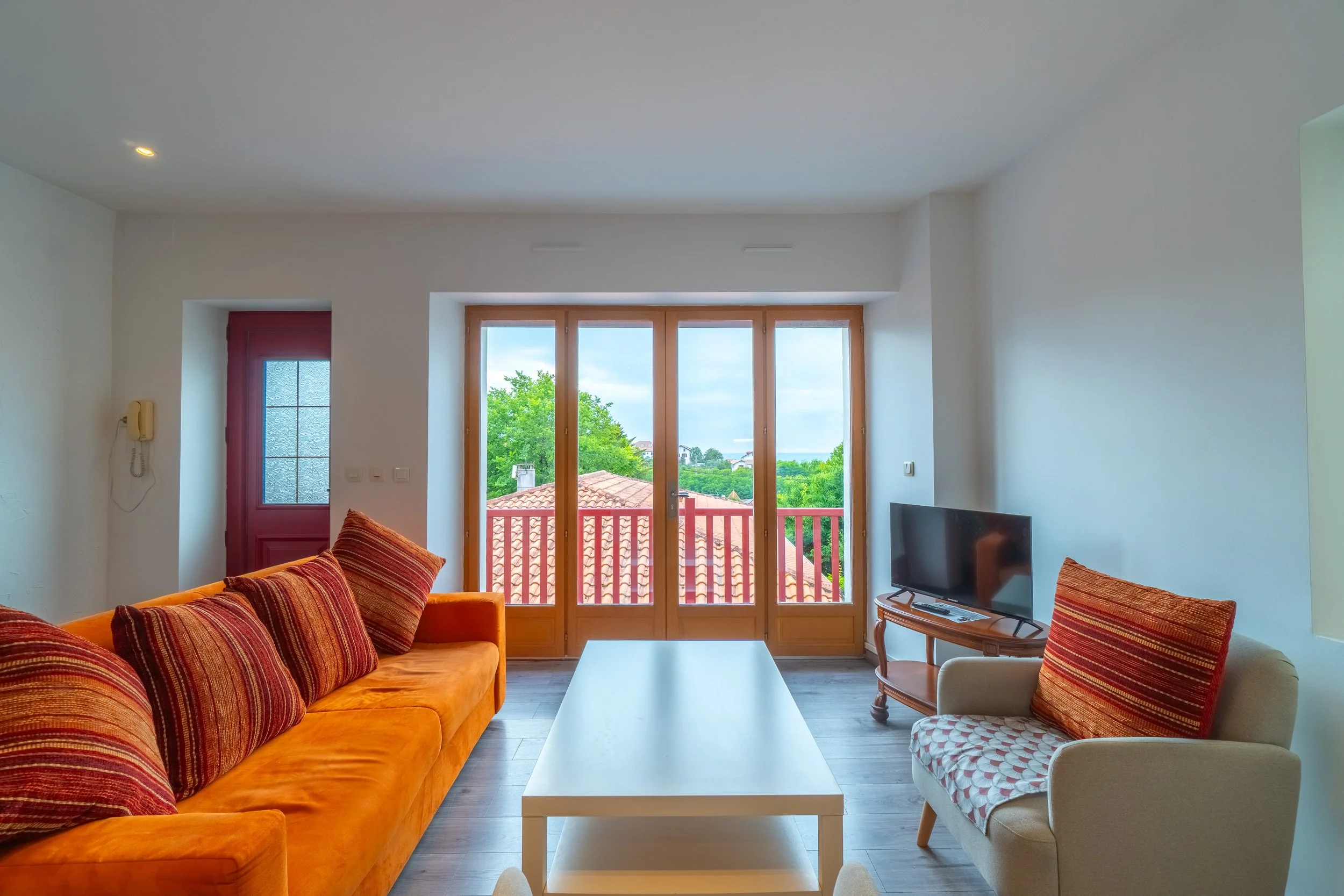 Salon avec canapé orange, fauteuil beige, table basse blanche, télévision, accès à un balcon avec vue sur des toits et des arbres, murs blancs, porte-fenêtre en bois.