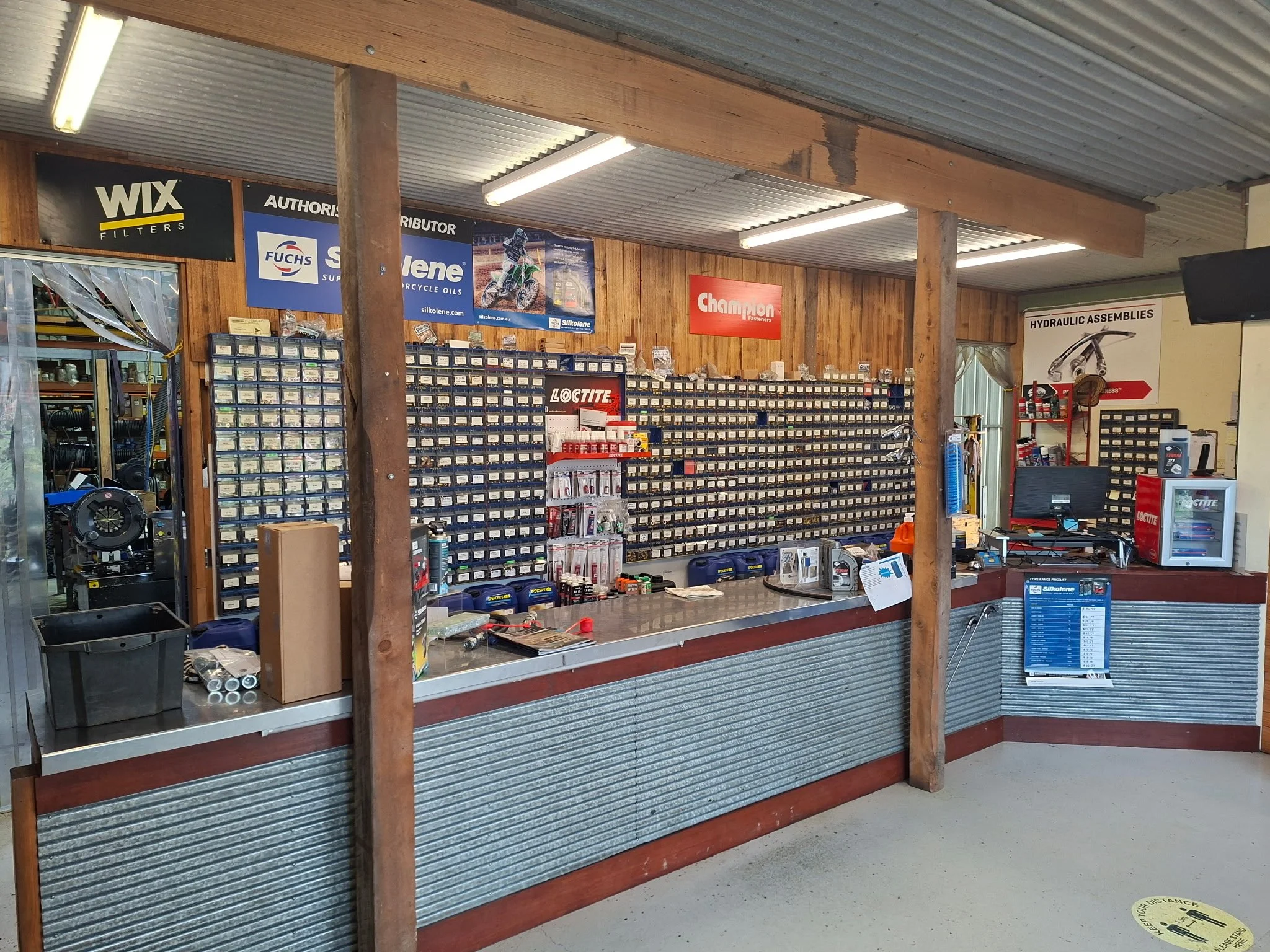 Inside a hardware or auto parts store with shelves filled with small labeled containers, signs from brands like Wix, Fuchs, Silkolene, and Loctite, and various tools and products on the counter.