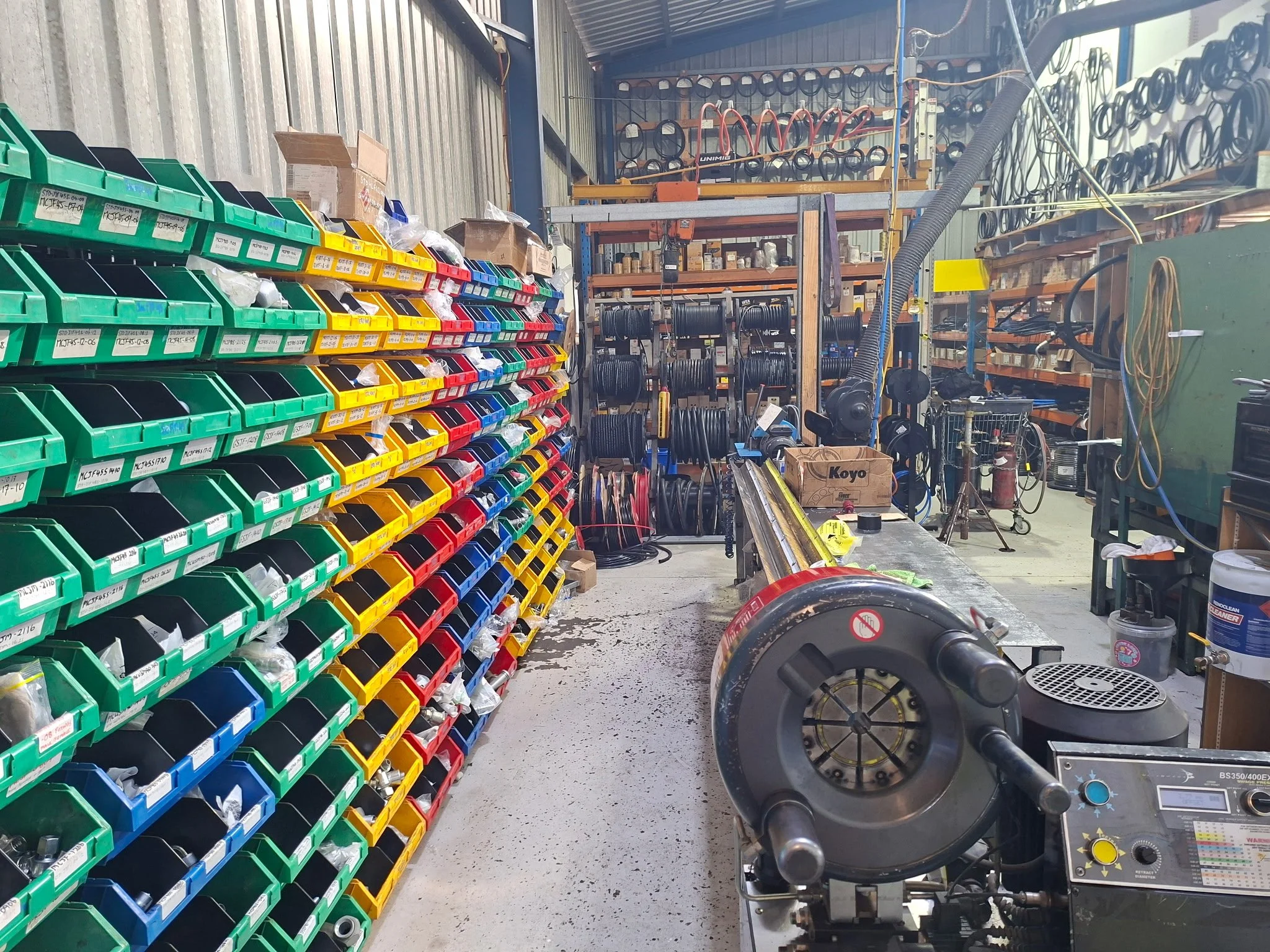 A workshop with organized shelves holding various parts, coils, and tools, including a stationary bike and a workbench with equipment.
