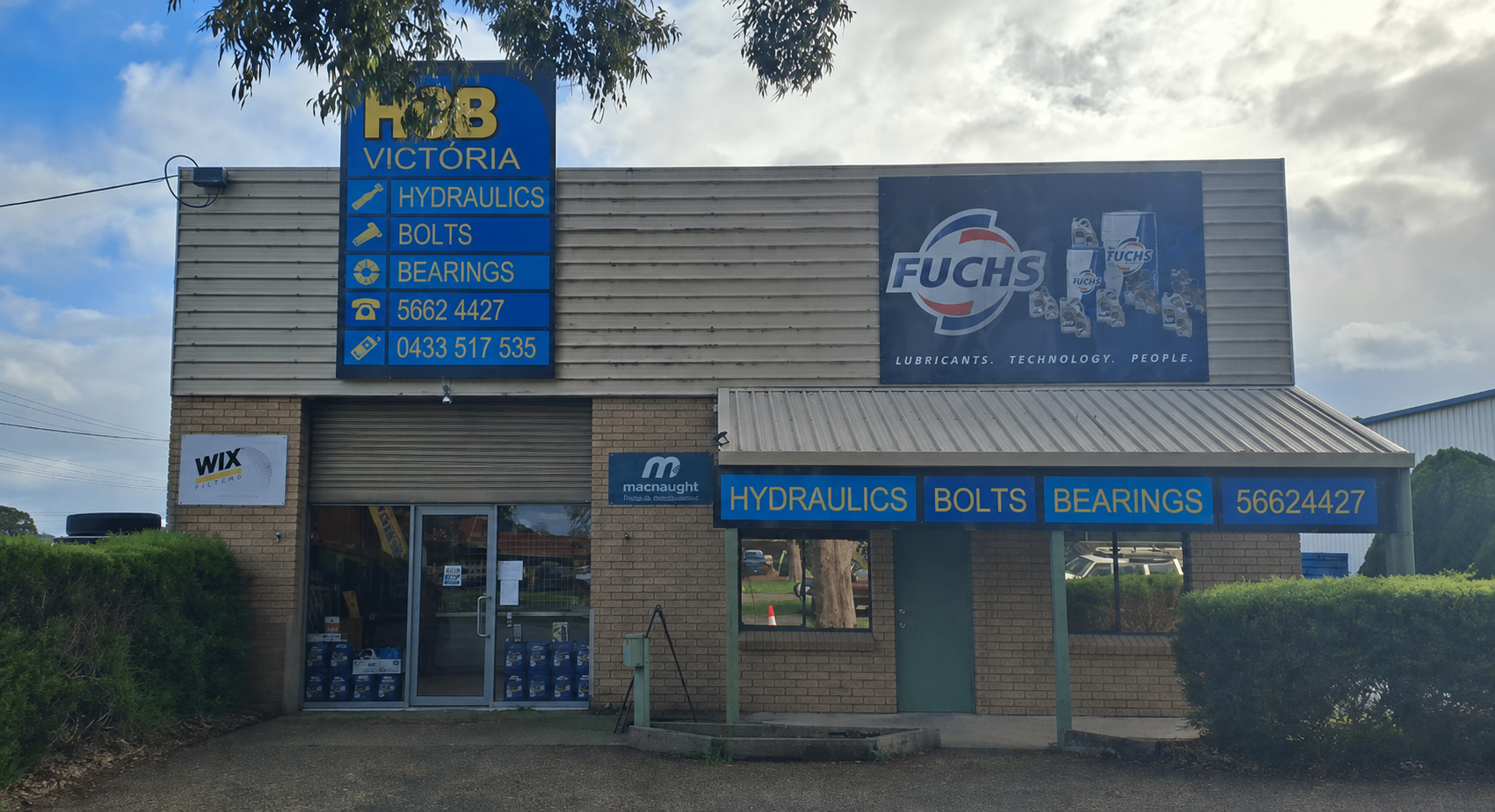 Exterior view of an automotive parts store with signs advertising hydraulics, bolts, bearings, and contact numbers, and a large FUCHS lubricant advertisement on the building.