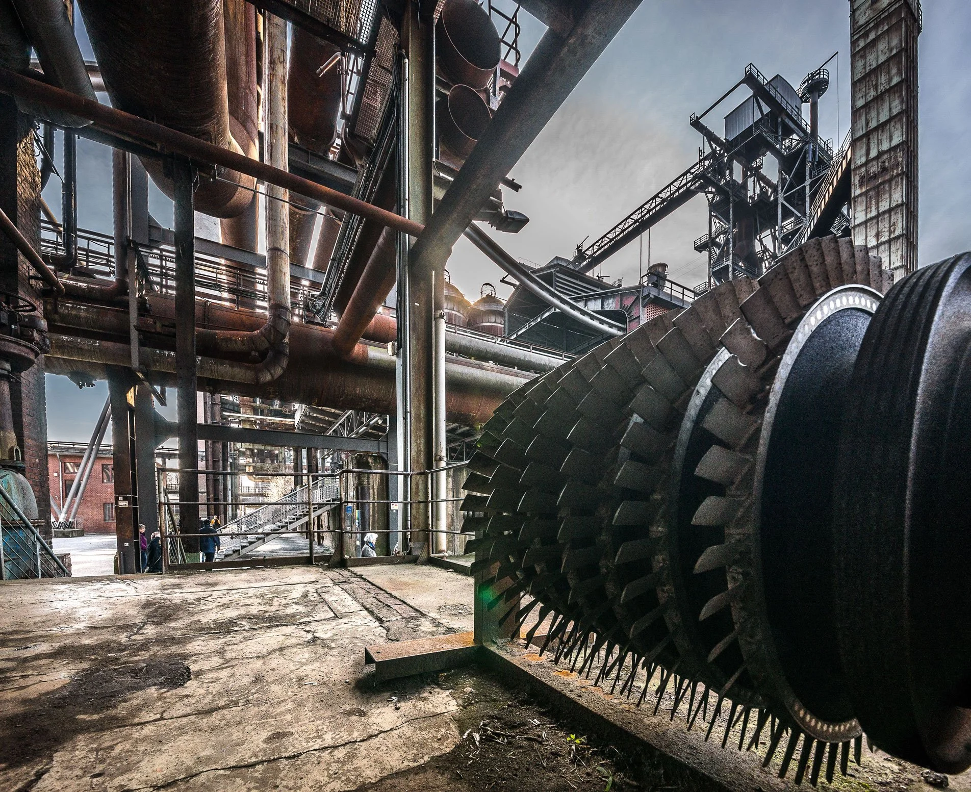 Industrial scene with old metal pipes, scaffolding, and machinery, viewed from inside a factory or plant with a cloudy sky in the background.