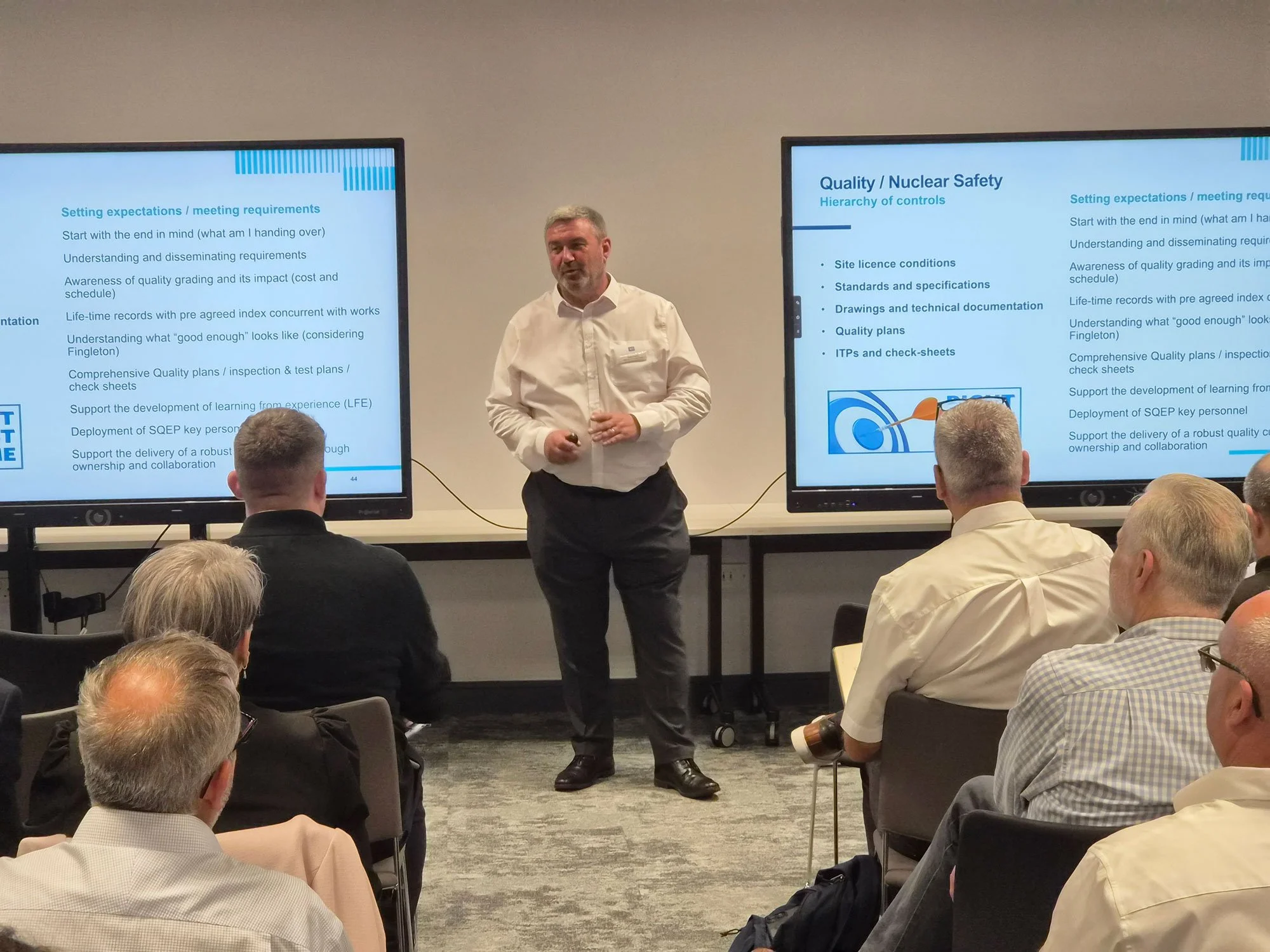 A man in a white shirt is giving a presentation in front of an audience. Two large screens display slides titled 'Quality / Nuclear Safety' with bullet points. The attendees are seated, facing the speaker and screens.