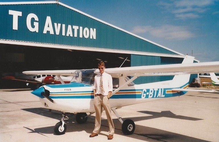 A man standing next to a small white and blue airplane in front of a hangar with 'T G AVIATION' written on it.