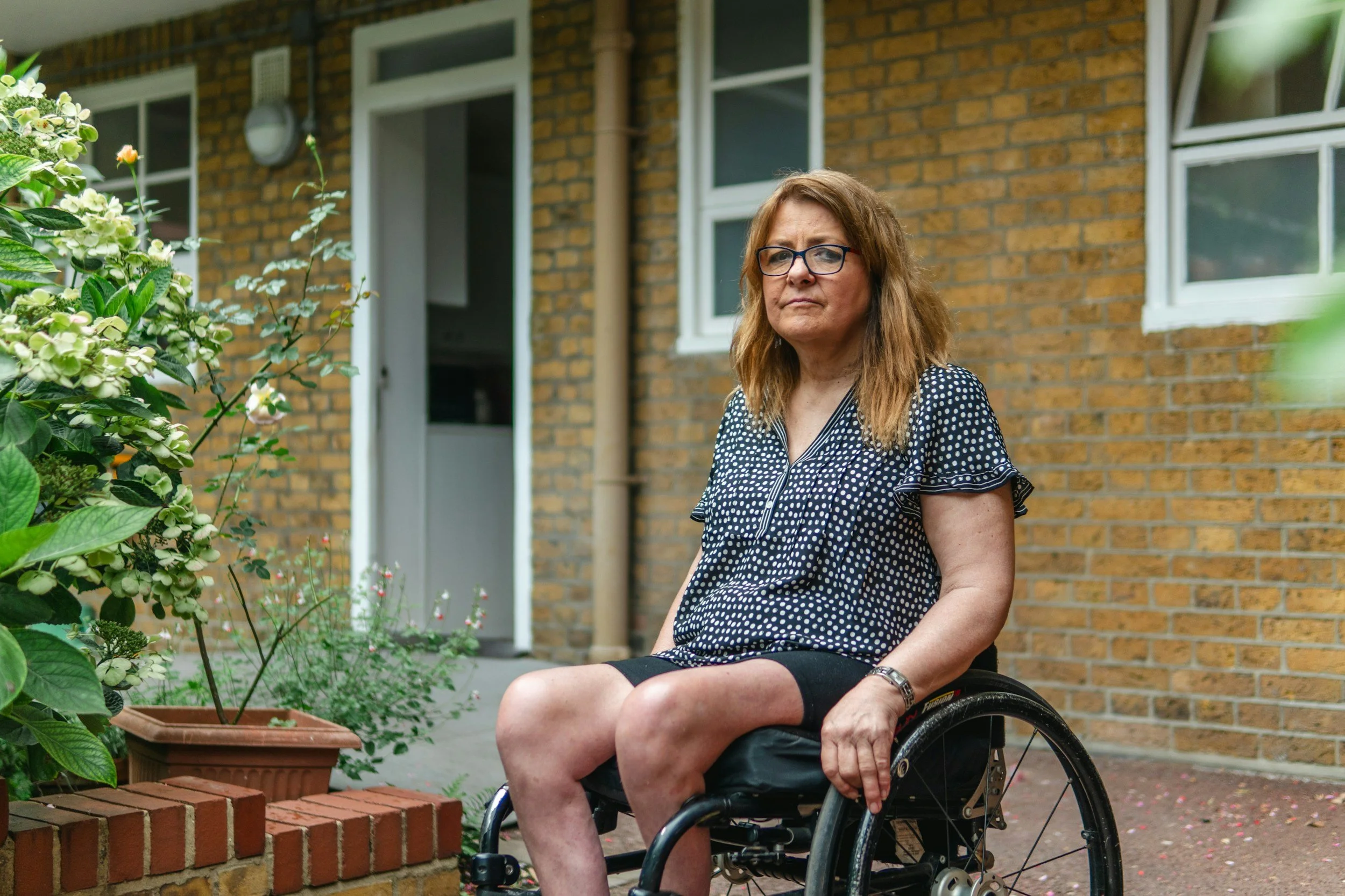 A woman with glasses and wavy reddish-brown hair sits in a wheelchair outside a brick house, surrounded by green plants and flowers.