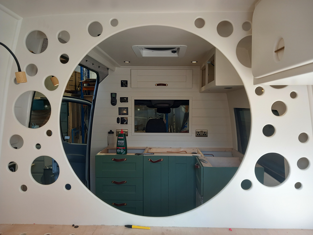 Interior view of a tiny house or caravan under construction, seen through a large circular opening with decorative holes. Inside, there are green cabinets, a window, and various construction tools and supplies.