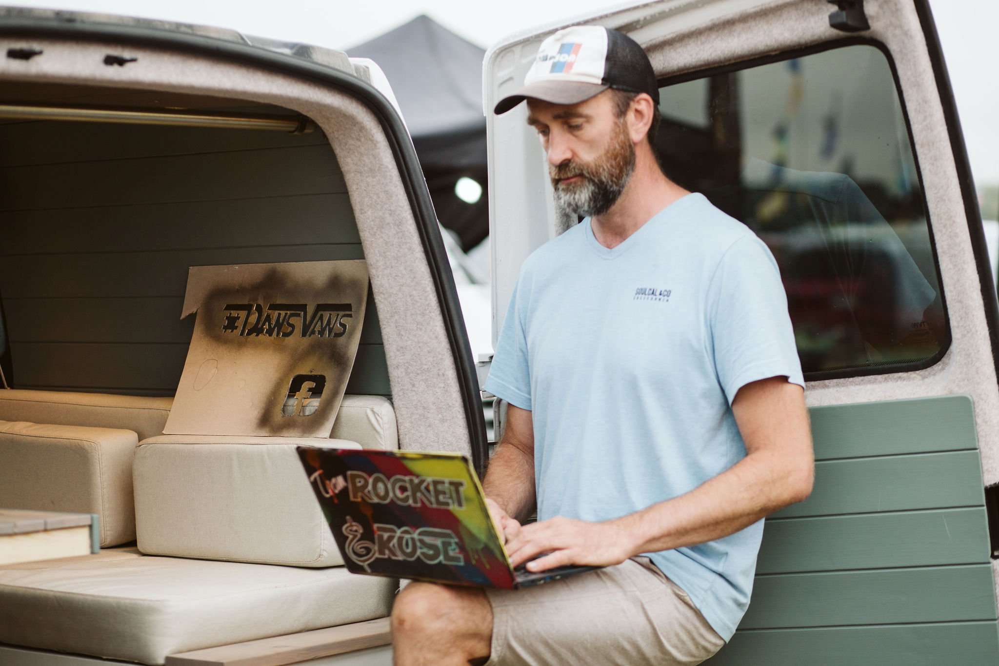 A man with a beard wearing a baseball cap and a light blue t-shirt, sitting in a camper van, looking at a laptop. The camper van's interior shows a sign that says '#DANSVANS' and a sticker on the window that says 'TROCKET'.