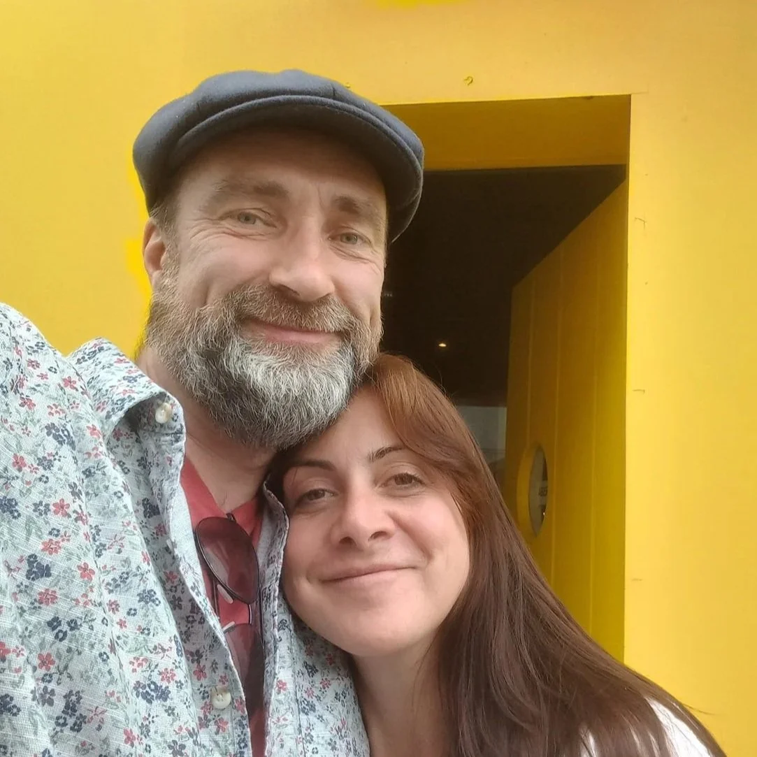 A smiling man with a gray beard, wearing a dark cap and a floral shirt, taking a close-up selfie with a woman with reddish-brown hair, smiling. They are standing in front of a yellow background.