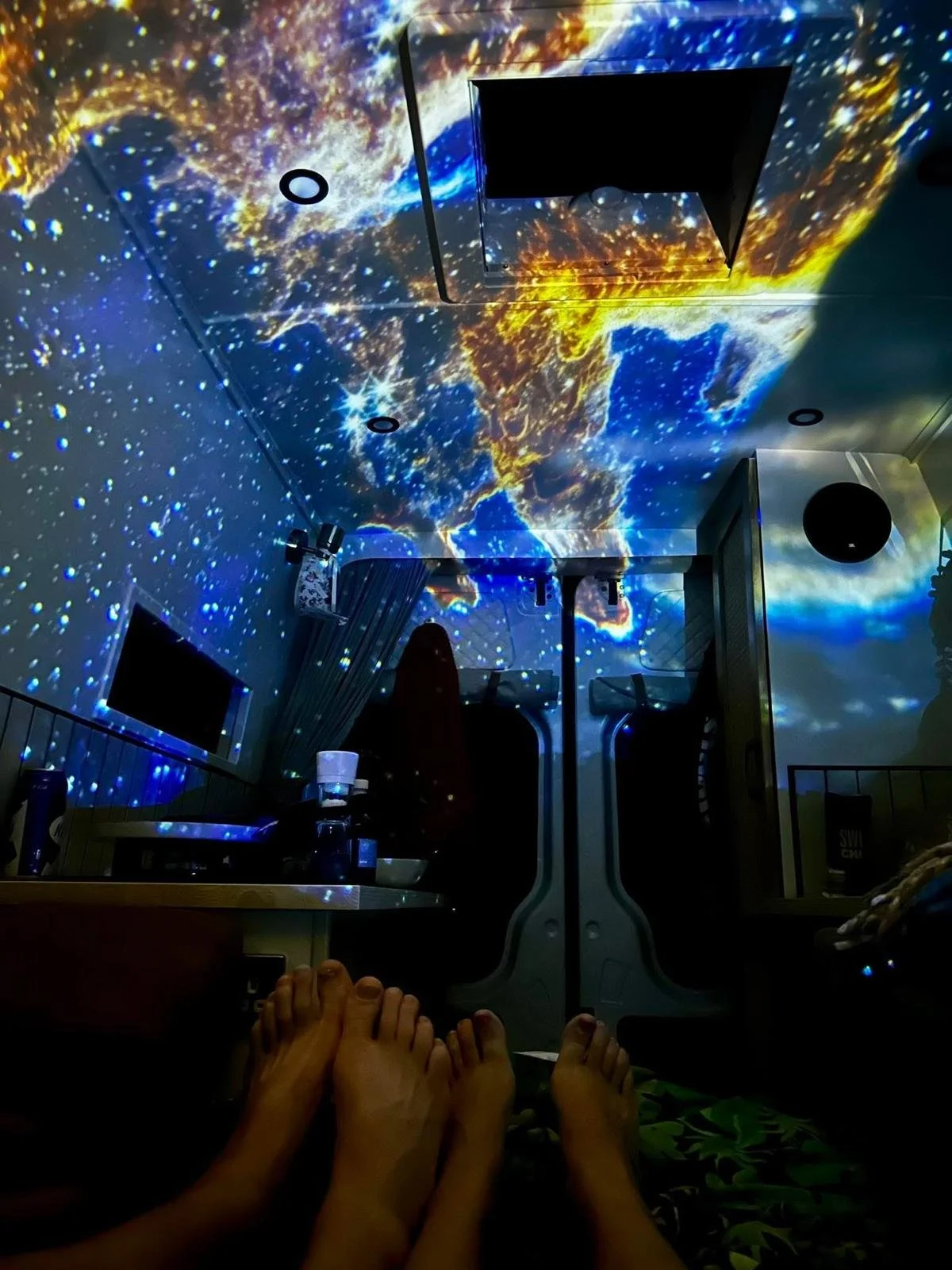 Room with a projection of Earth from space on the ceiling and walls, showing continents and clouds, creating a space-themed atmosphere. There are people relaxing with their feet visible, a table with bottles and cups, and monitors mounted on the walls.