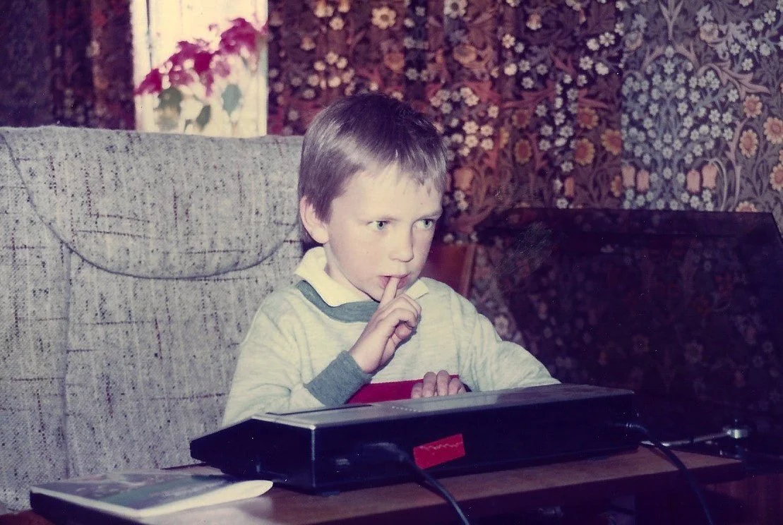 A young boy with short light brown hair sitting at a table, looking at a laptop with a finger pressed to his lips, in a room with floral wallpaper and a floral curtain.