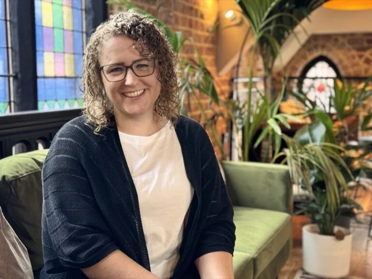 A woman with curly hair, glasses, and a white shirt sitting on a green couch in a warmly lit indoor space with plants and stained glass windows.